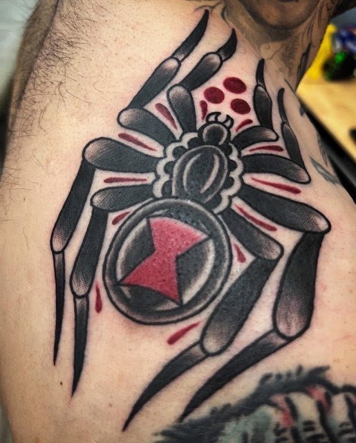 Tattoo of a spider with a pocket watch on its body, surrounded by red dots and streaks on a person's shoulder.