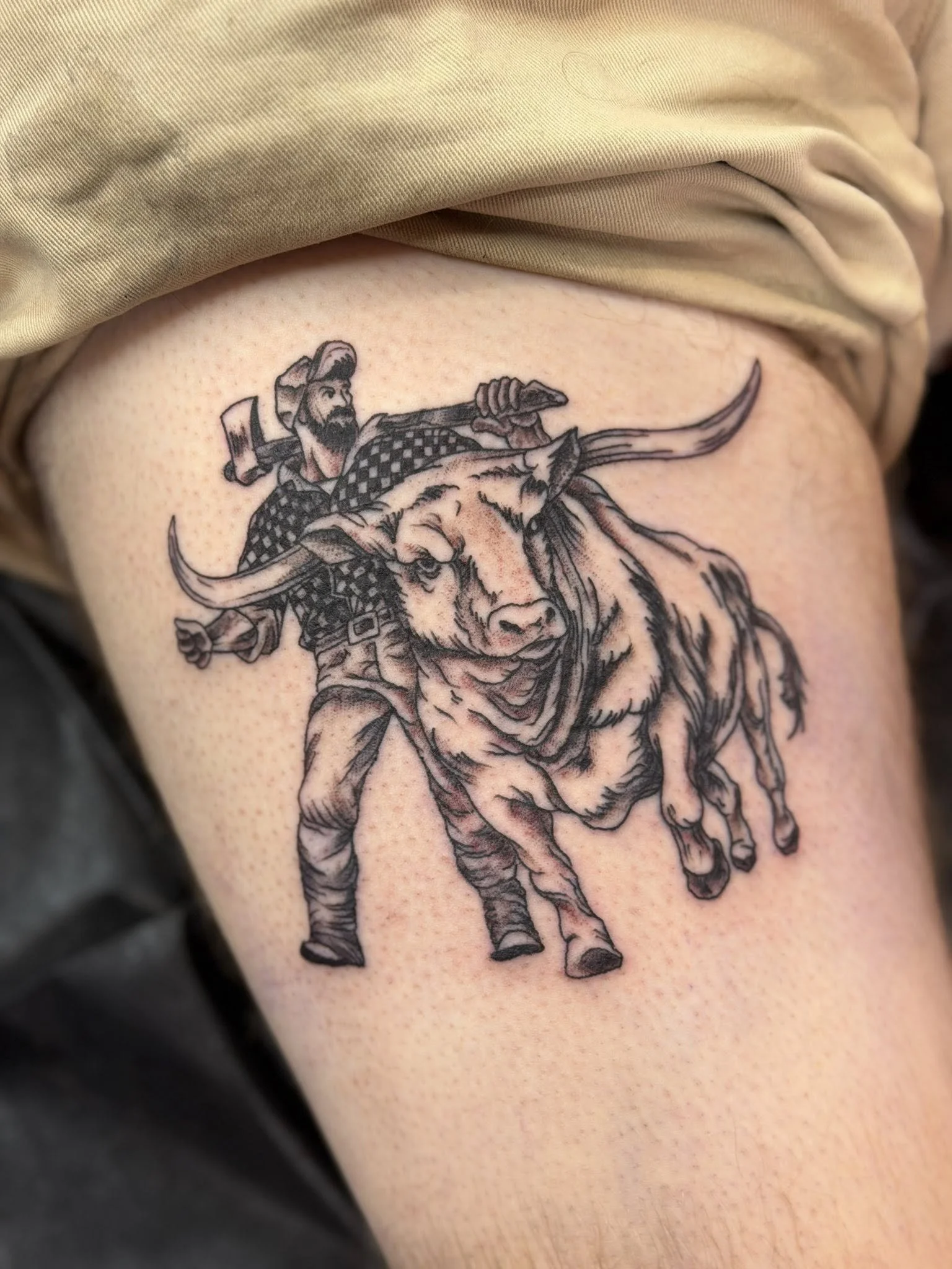 Tattoo of a cowboy with a beard and checkered shirt riding a large bull with long horns.