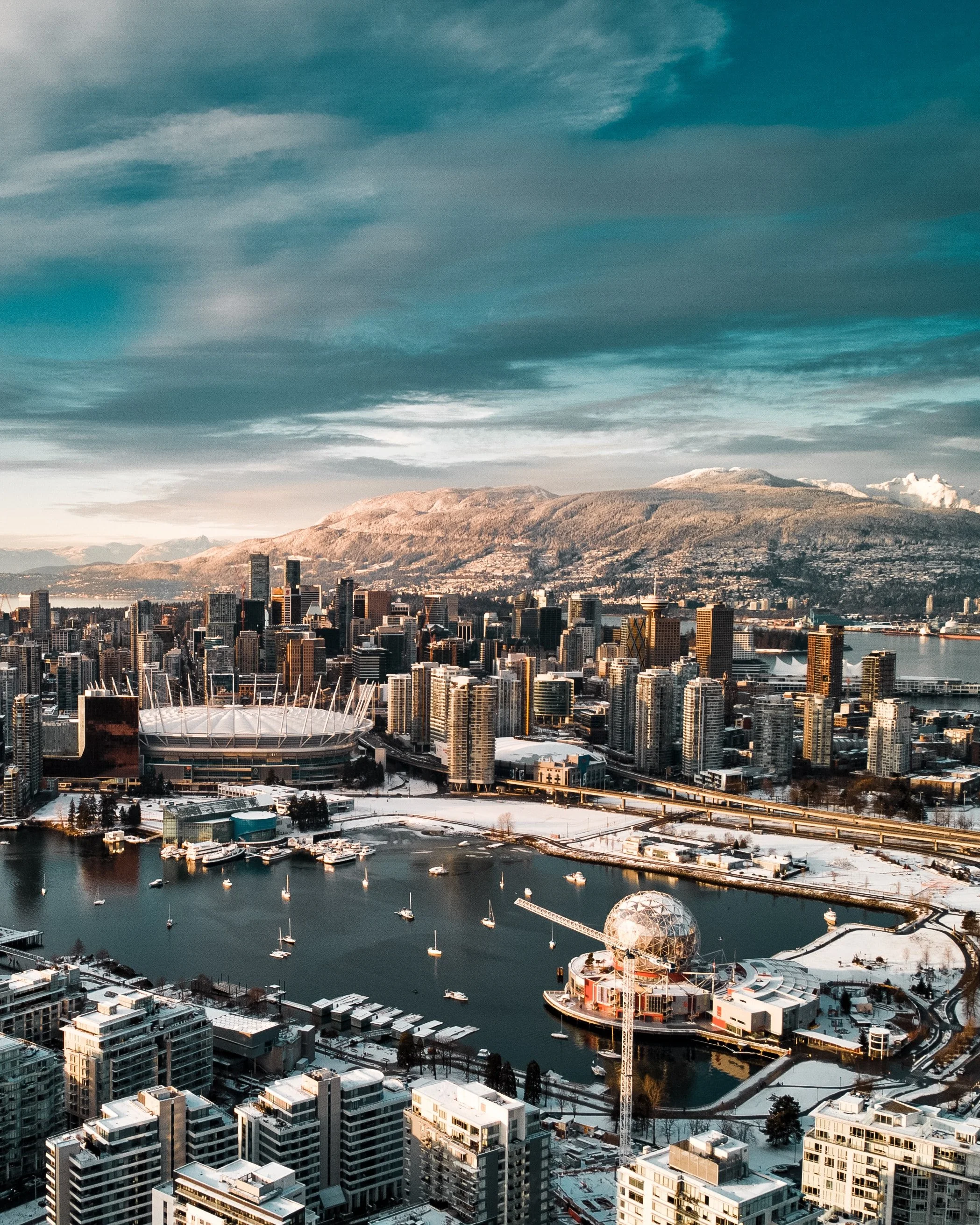 Aerial view of Vancouver, Canada, with city skyscrapers, a snow-covered landscape, a harbor with boats, and mountains in the background under a partly cloudy sky.