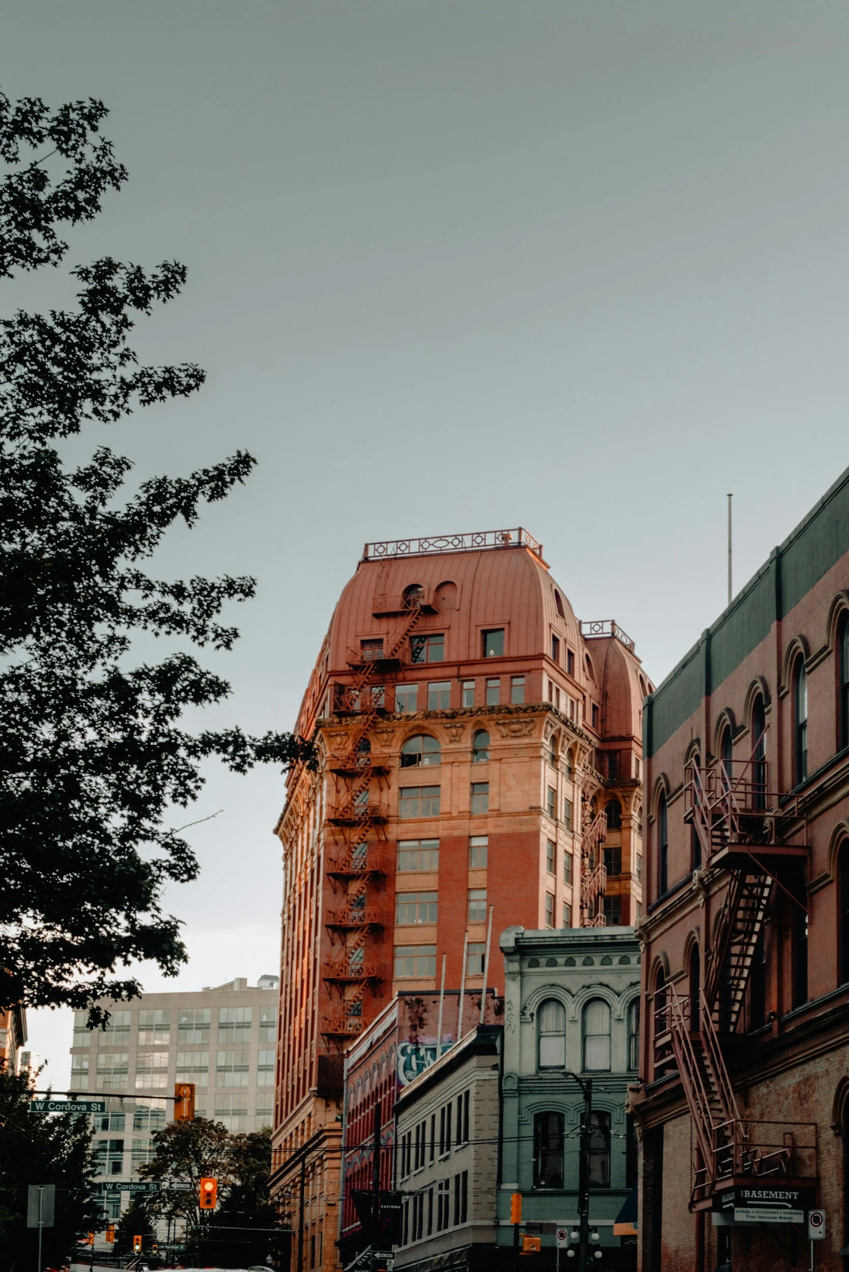 Tall historical building with fire escape stairs on the outside, located on a city street with traffic lights and other buildings.