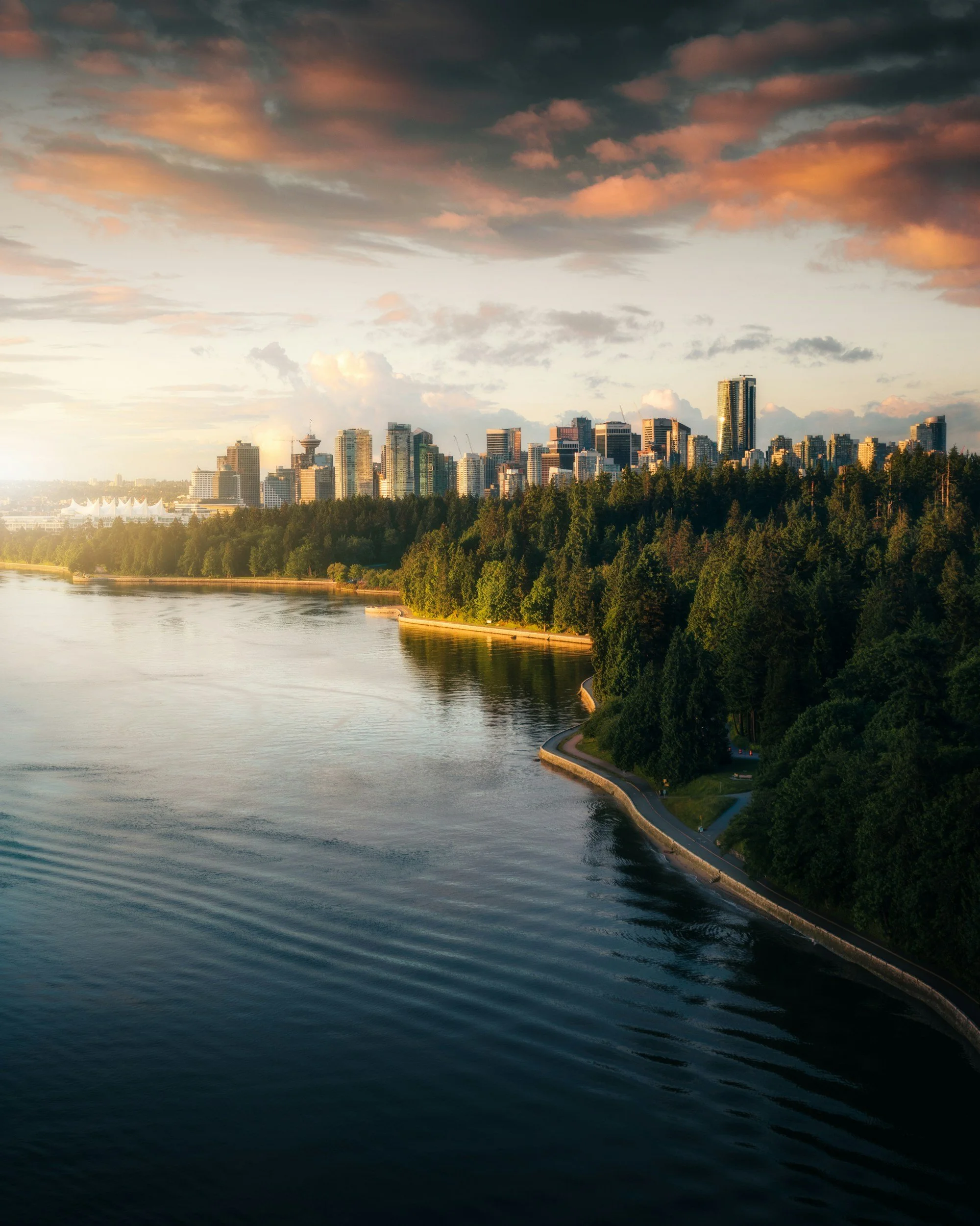 A city skyline at sunset with tall buildings, a body of water in the foreground, and a tree-lined shoreline.