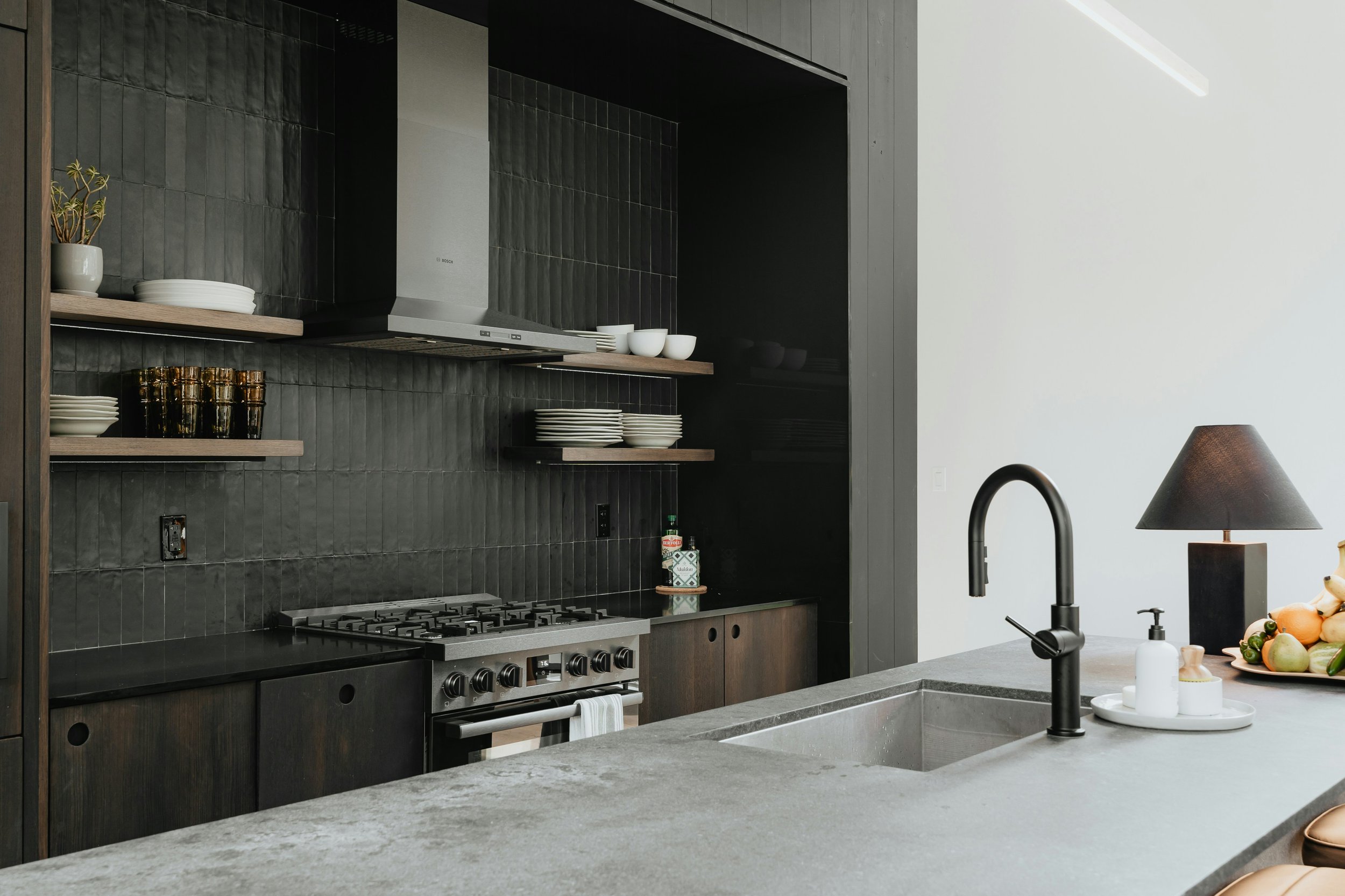 Modern kitchen with black tiled backsplash, open shelving with white and black dishes, a stainless steel stove, a black faucet over gray sink, and a gray island with a lamp, soap dispenser, and fruit on it.