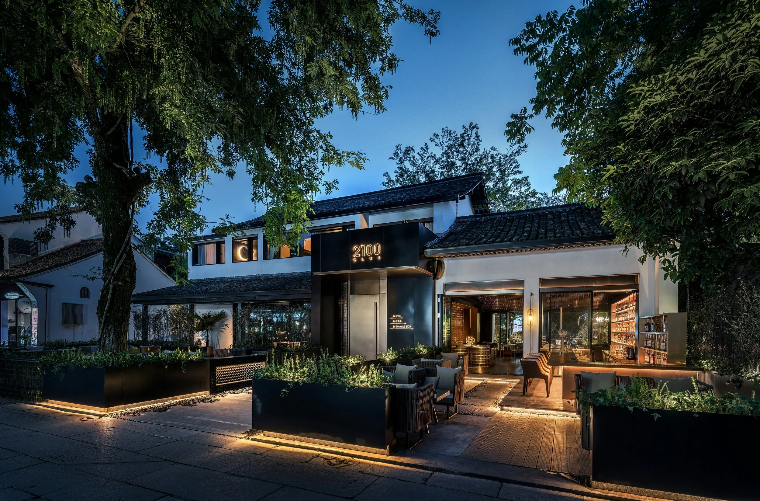 Exterior of a modern restaurant or cafe with outdoor seating, illuminated signs, and lush trees at dusk.