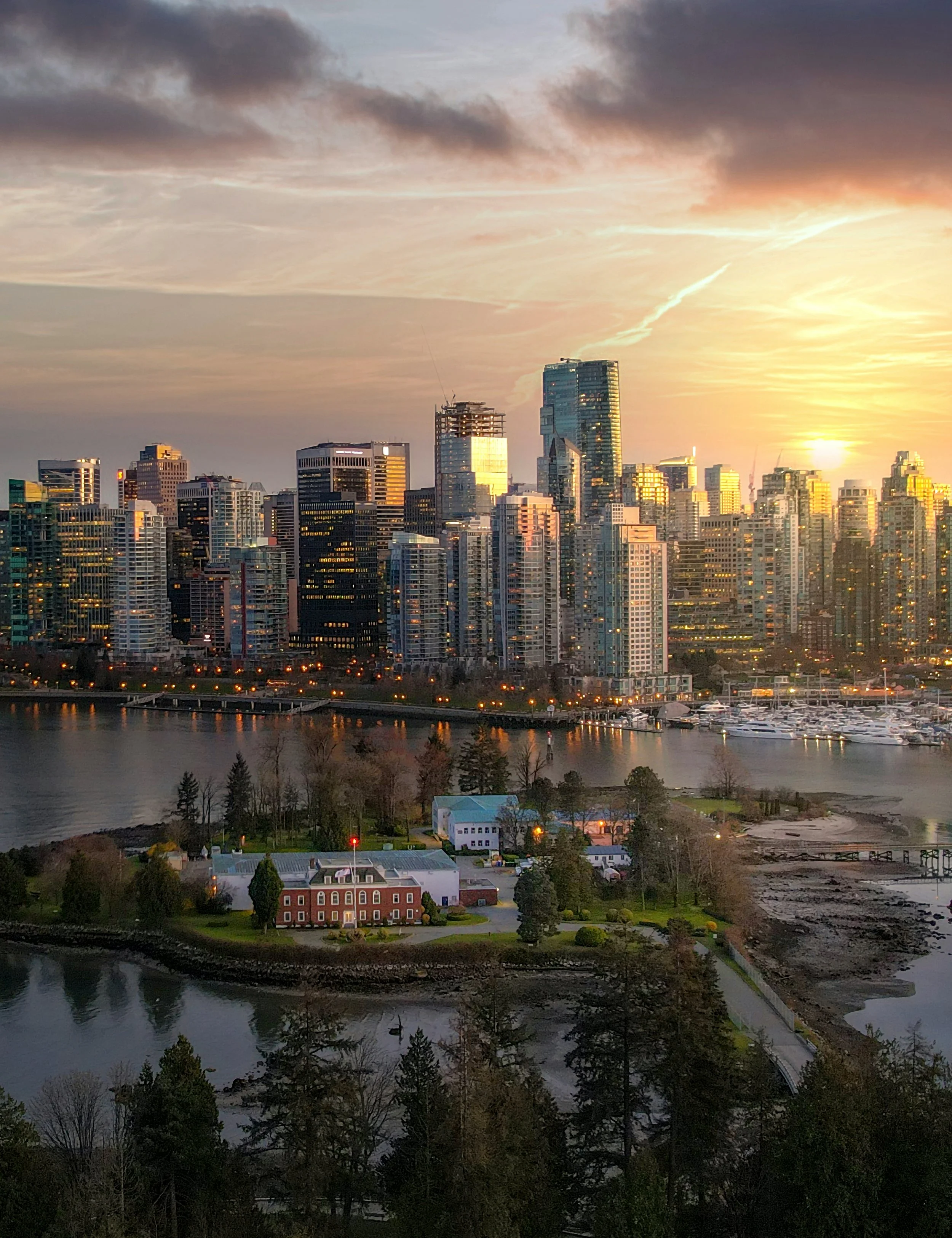 City skyline at sunset with tall skyscrapers, a river reflecting the buildings, a small historic brick building on the waterfront, and a marina with boats.