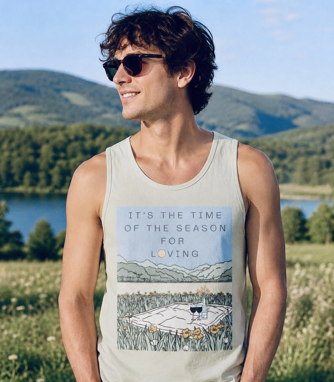 Man wearing sunglasses and a sleeveless shirt with a scenic graphic that says 'It's the time of the season for loving', standing outdoors in a field with mountains and a lake in the background.