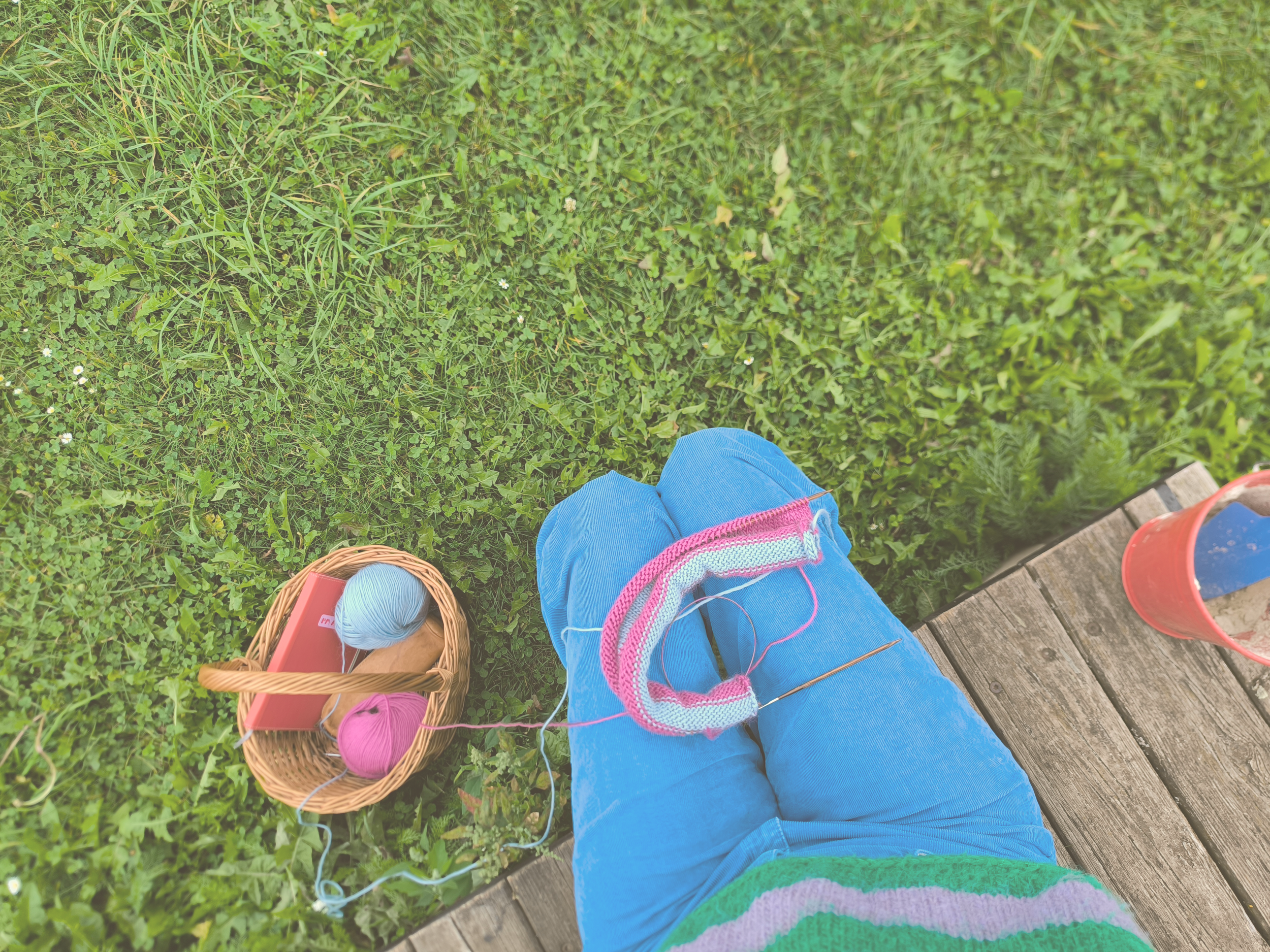 Blick auf die Beine einer Person auf einer Gartenlaube, umgeben von grünem Gras, mit einem Korb, Wolle und Stricknadeln in der Nähe.