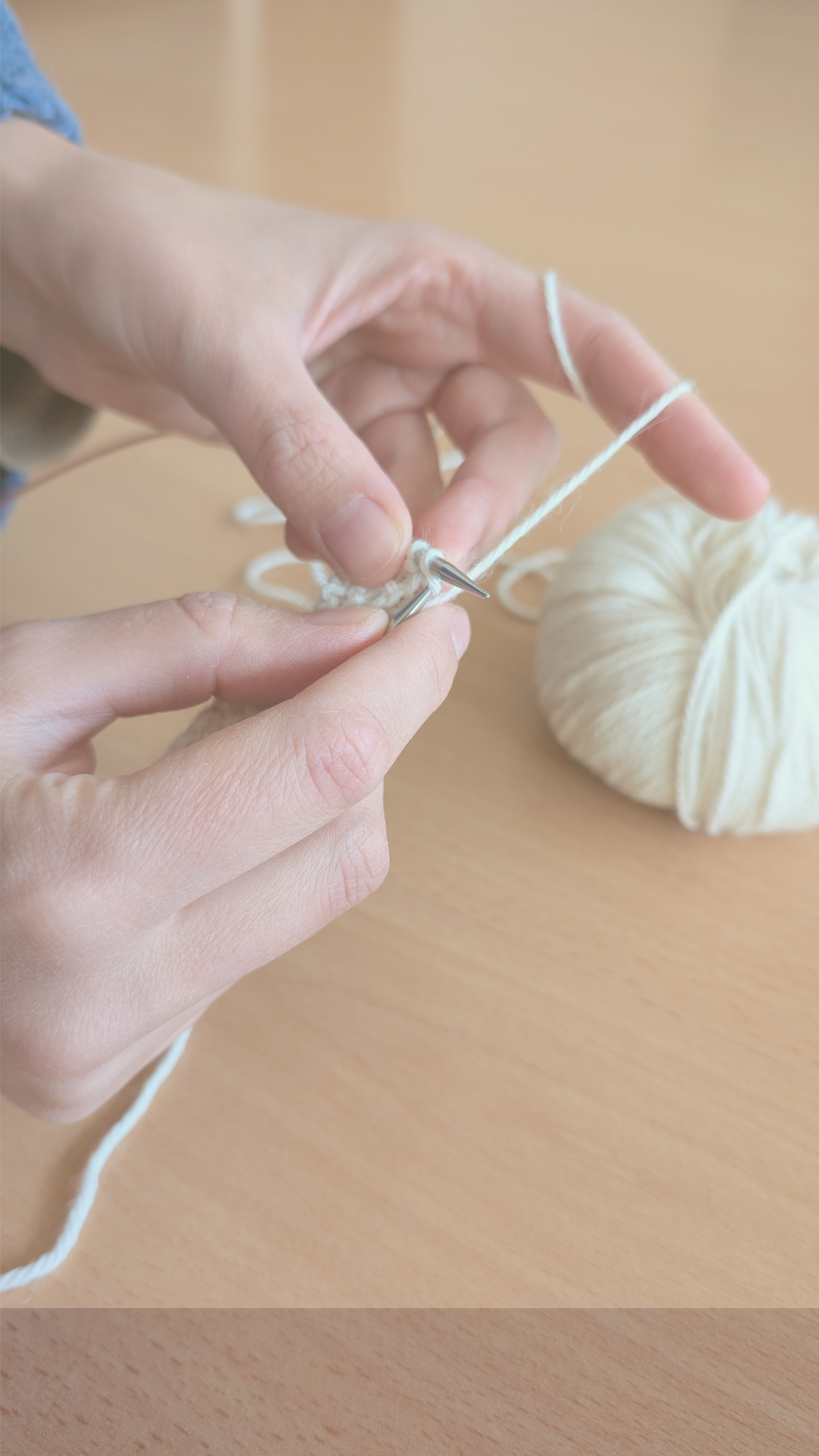 Nahaufnahme von Händen, die mit einer Nadel und weißer Wolle stricken, auf einem Holztisch.