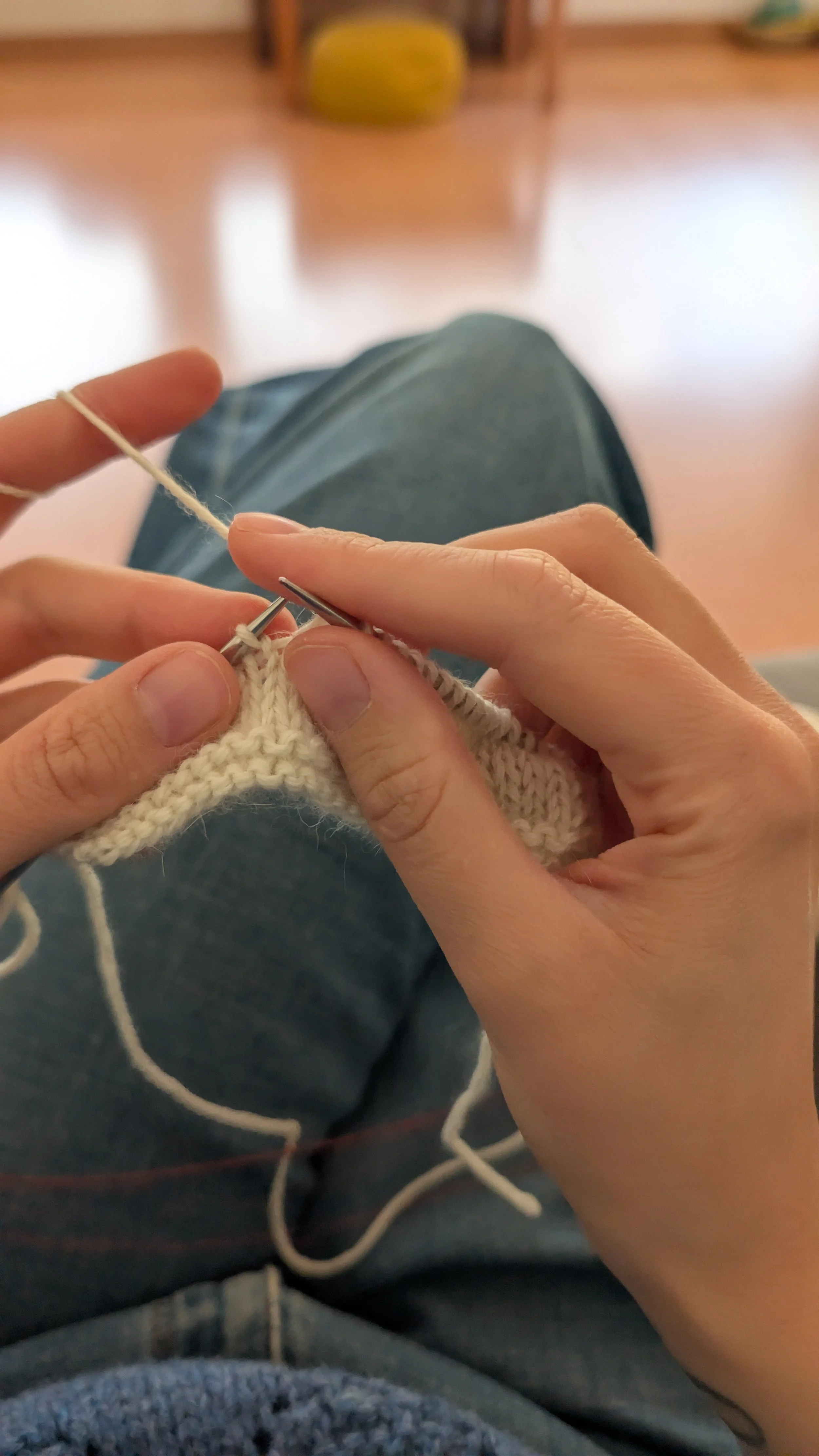 Eine Person sitzt mit gekreuzten Beinen und strickt mit weißen Garn und Nadeln in einem Innenraum, im Hintergrund ist ein gelber Sitzhocker sichtbar.