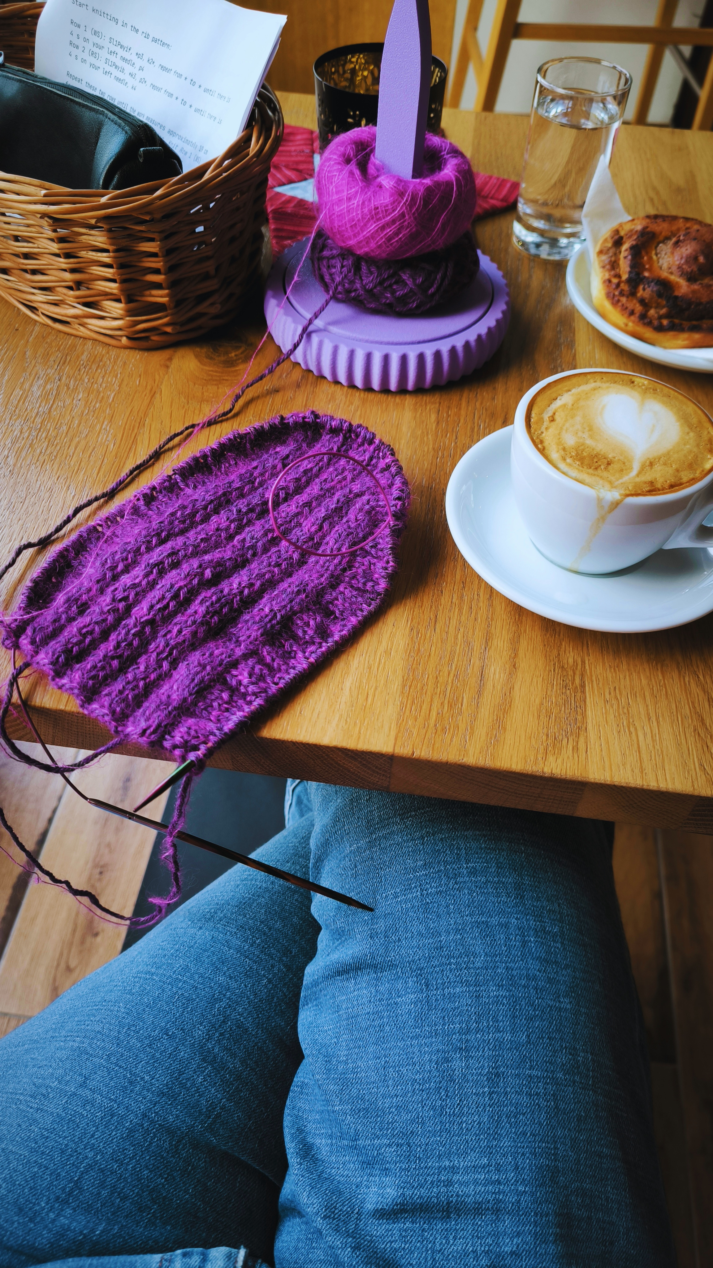 Person sitzt an einem Holztisch mit einer Tasse Kaffee, einem Glas Wasser, einem gebrauchtem Backwerk, einer Nadel mit pinker Wolle, einem Wollknäuel, einer Pflanze, einem Korb mit Dokumenten und einer Tasche.