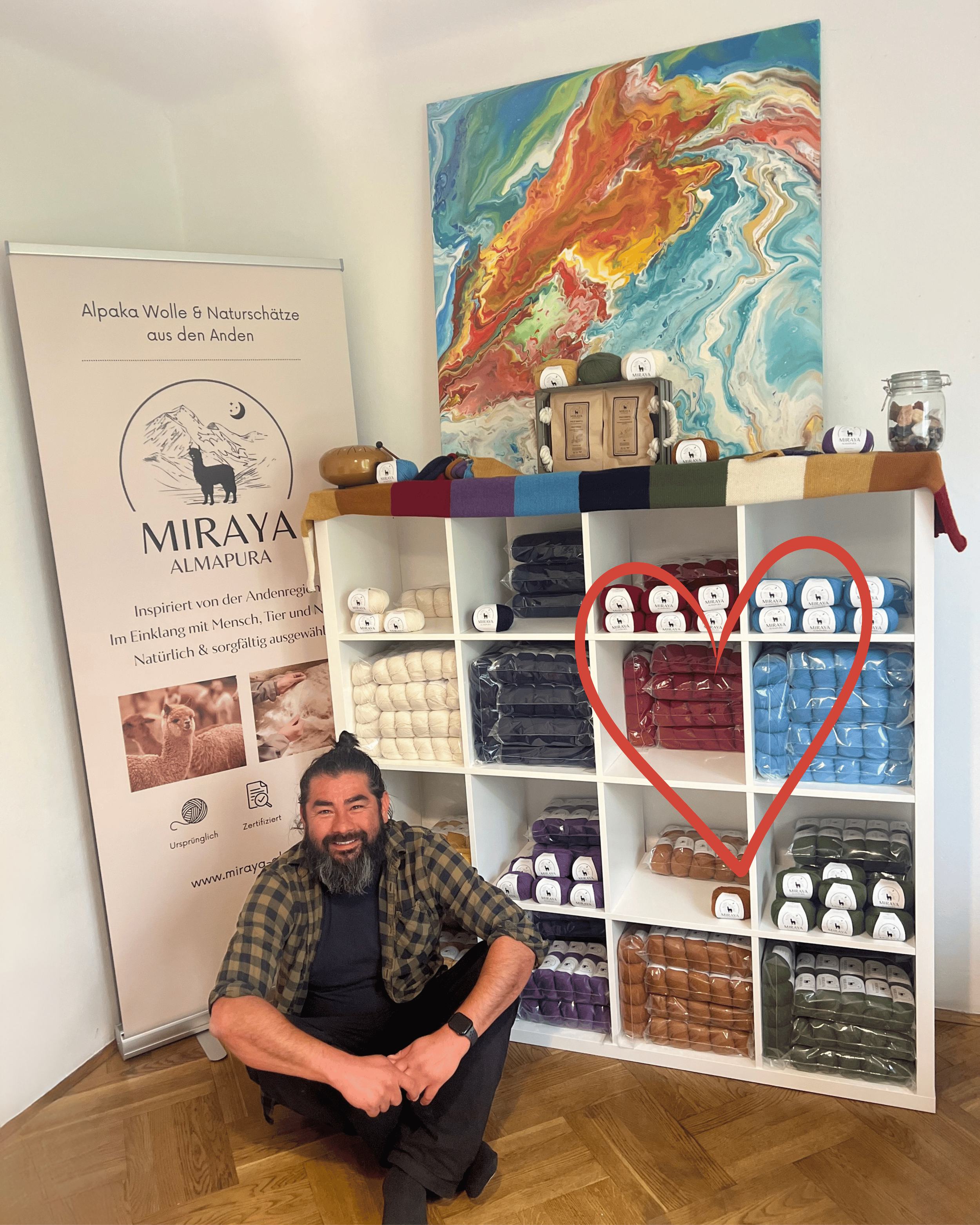 Der Gründer von Miraya Almapura sitzt vor einem Regal voller bunter Wolle aus 100% Babyalpaka im Büro von Miraya Almapura. Neben ihm eine Tafel mit Text über Alpaka Wolle und Naturmaterialien aus den Anden.