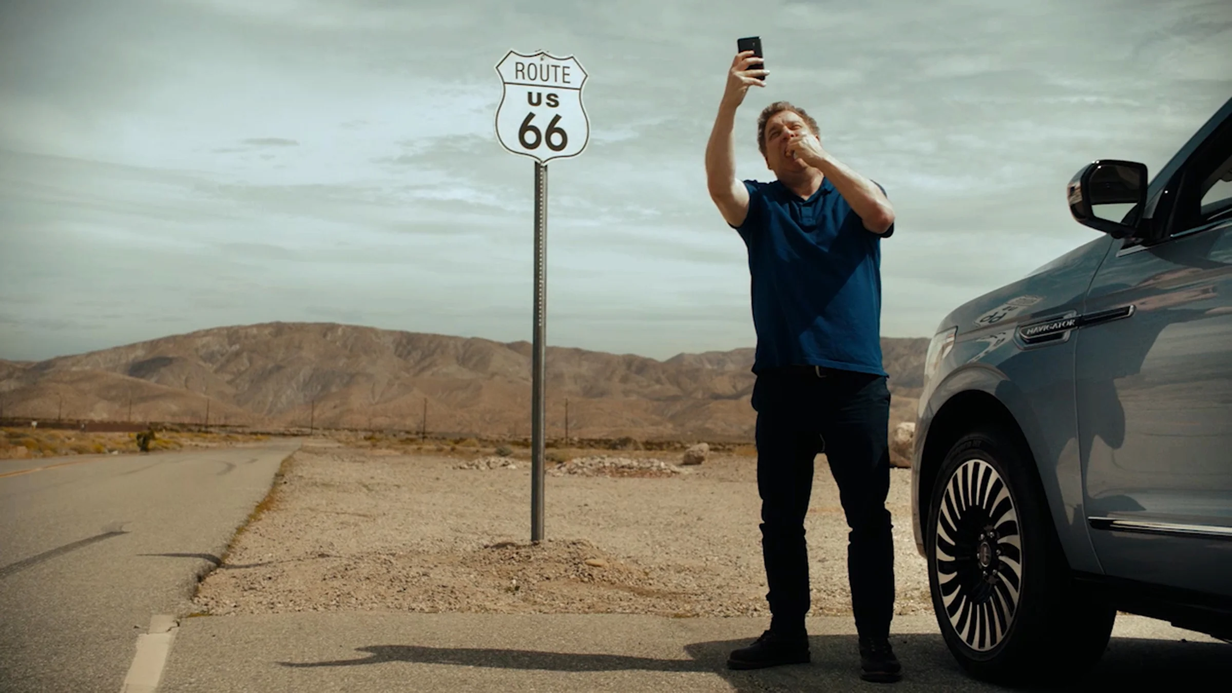 A man stands beside a gray car along the side of a desert highway, taking a selfie with a Route 66 sign and mountain landscape in the background.