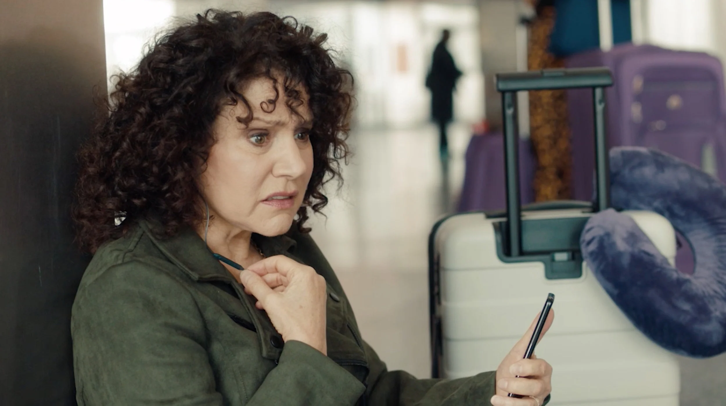 A woman with curly brown hair looks surprised or confused while looking at her phone, sitting at an airport with luggage nearby.