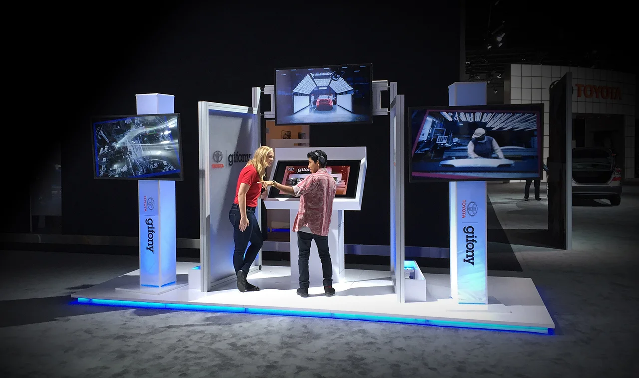 Two women exchanging a card or phone at an exhibition booth with Toyota and Gfory logos, surrounded by three large screens displaying automotive images.