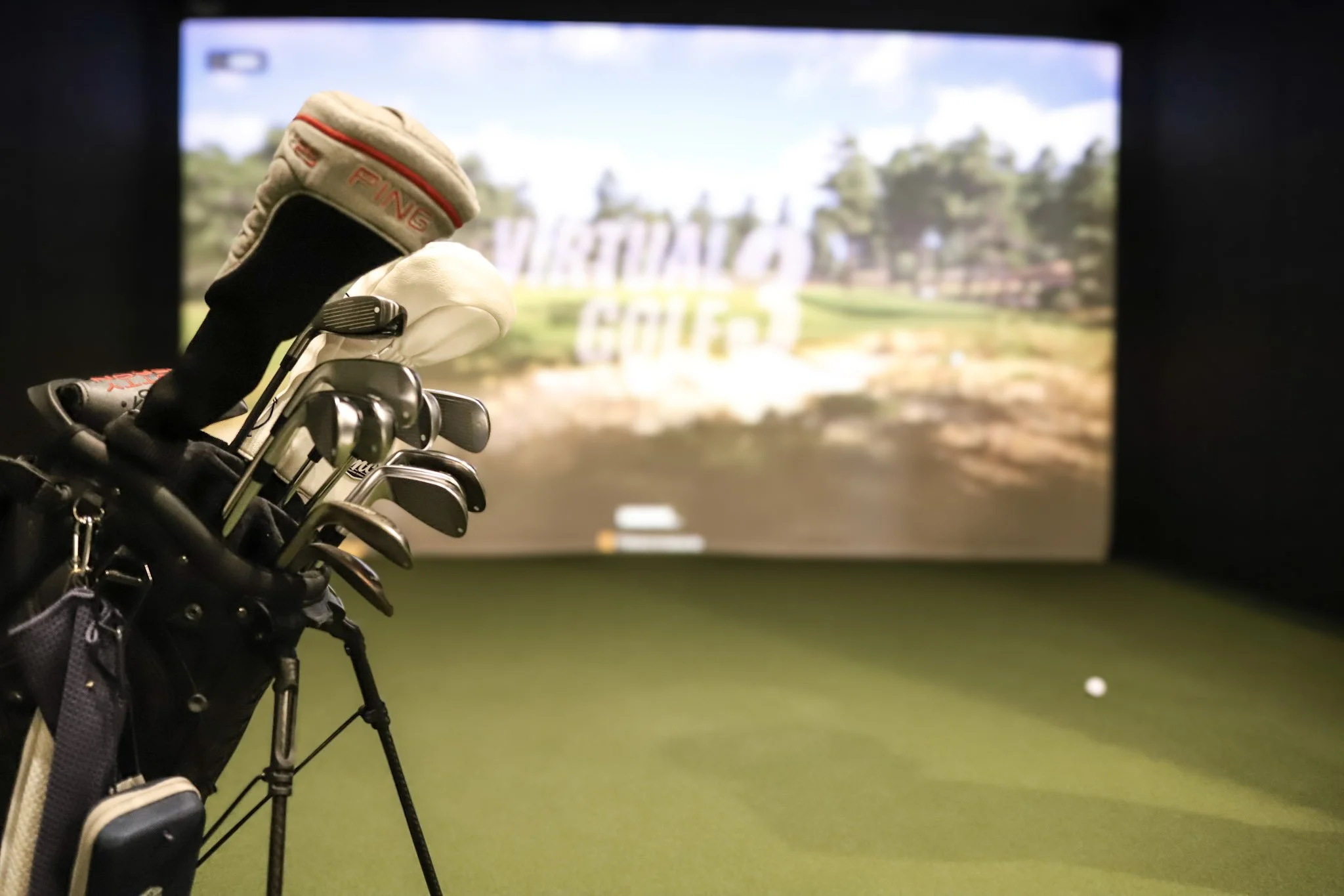 View of golf clubs arranged in a golf bag facing an indoor golf practice simulator with a golf course projection on the screen.
