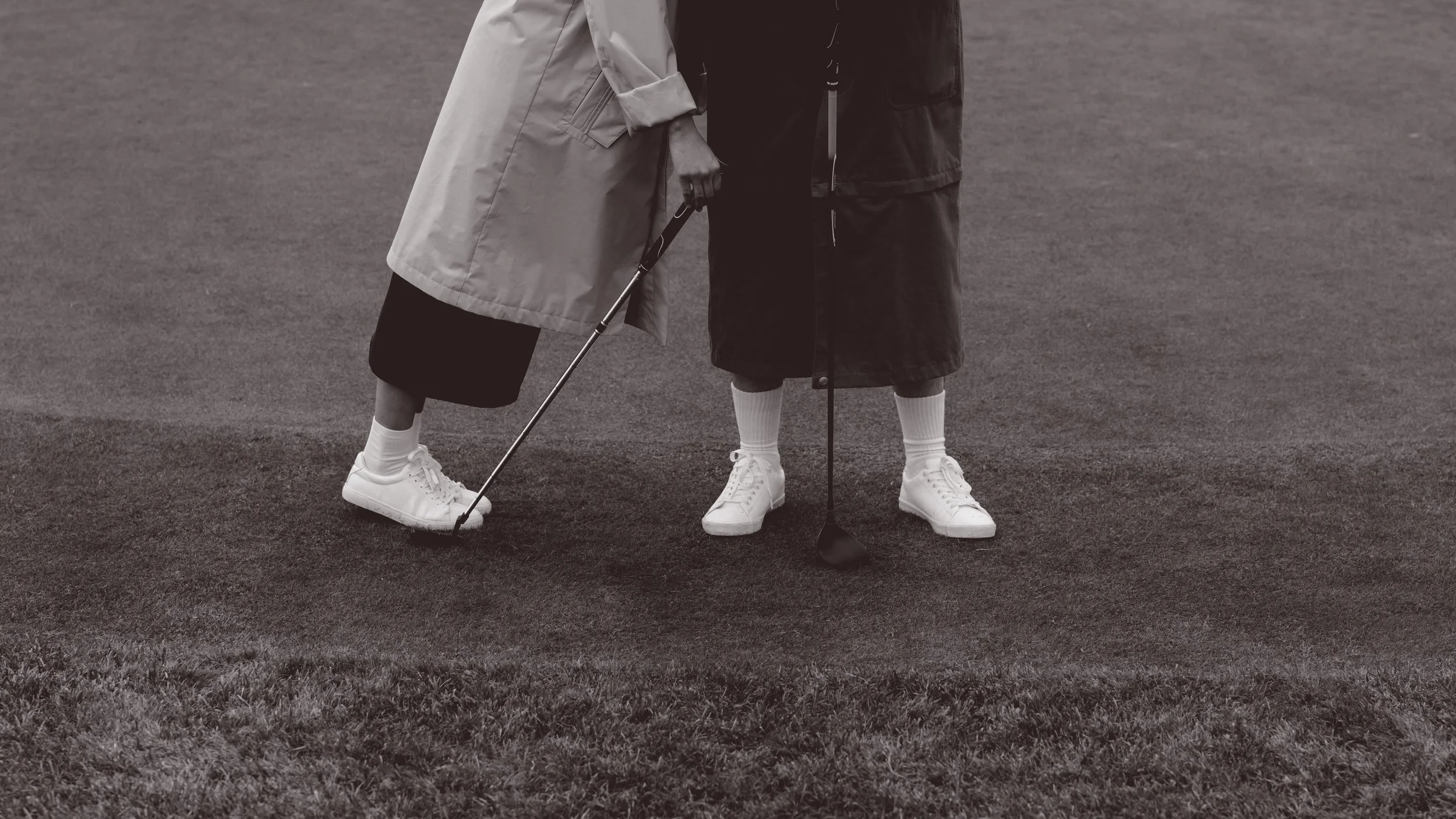 Two people are standing on a golf course putting with their golf clubs. One is wearing a light coat and the other a dark coat, both wearing white sneakers and socks.