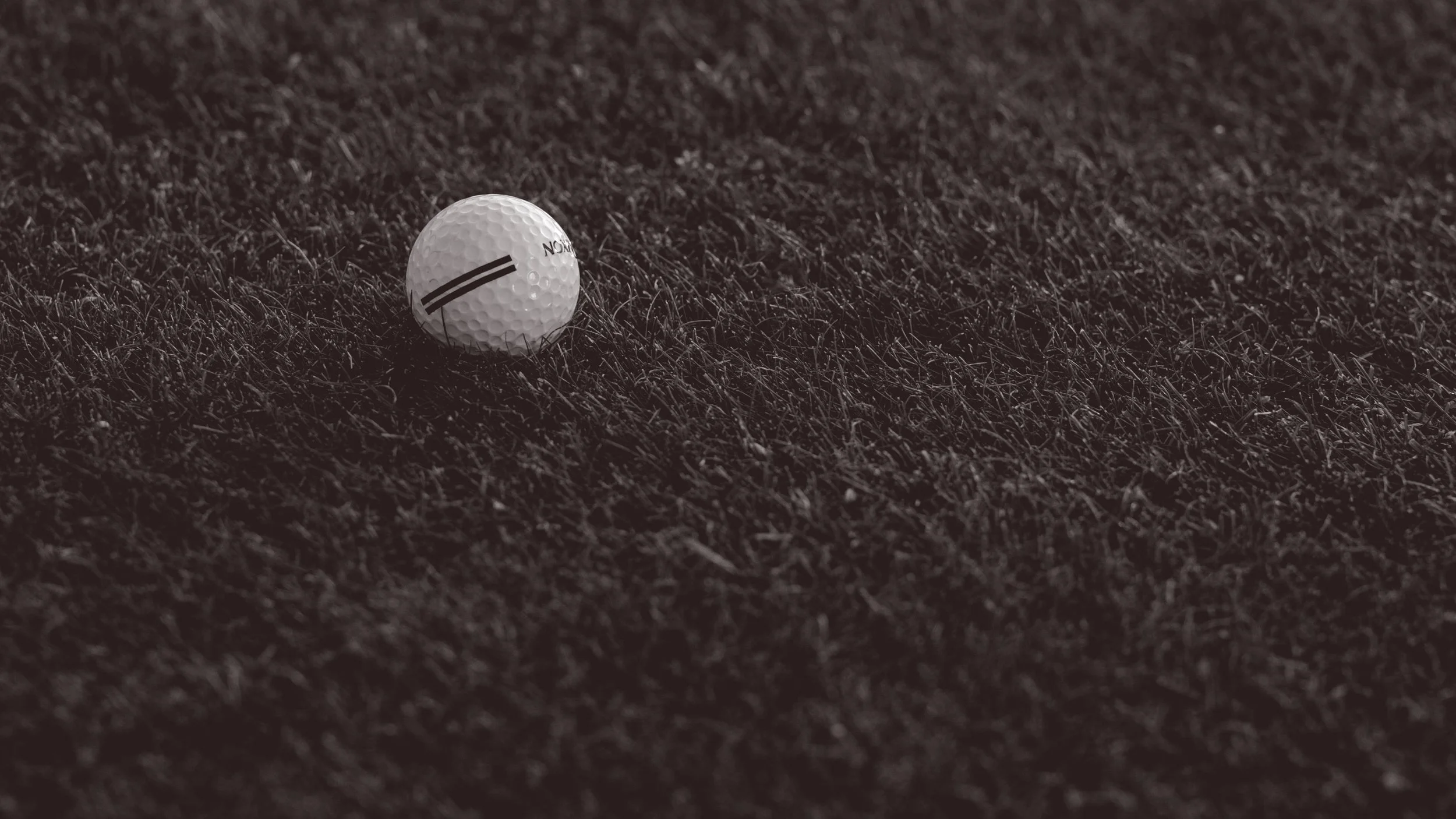 A close-up image of a golf ball on a dark, textured surface, possibly artificial turf, with the ball's markings visible.