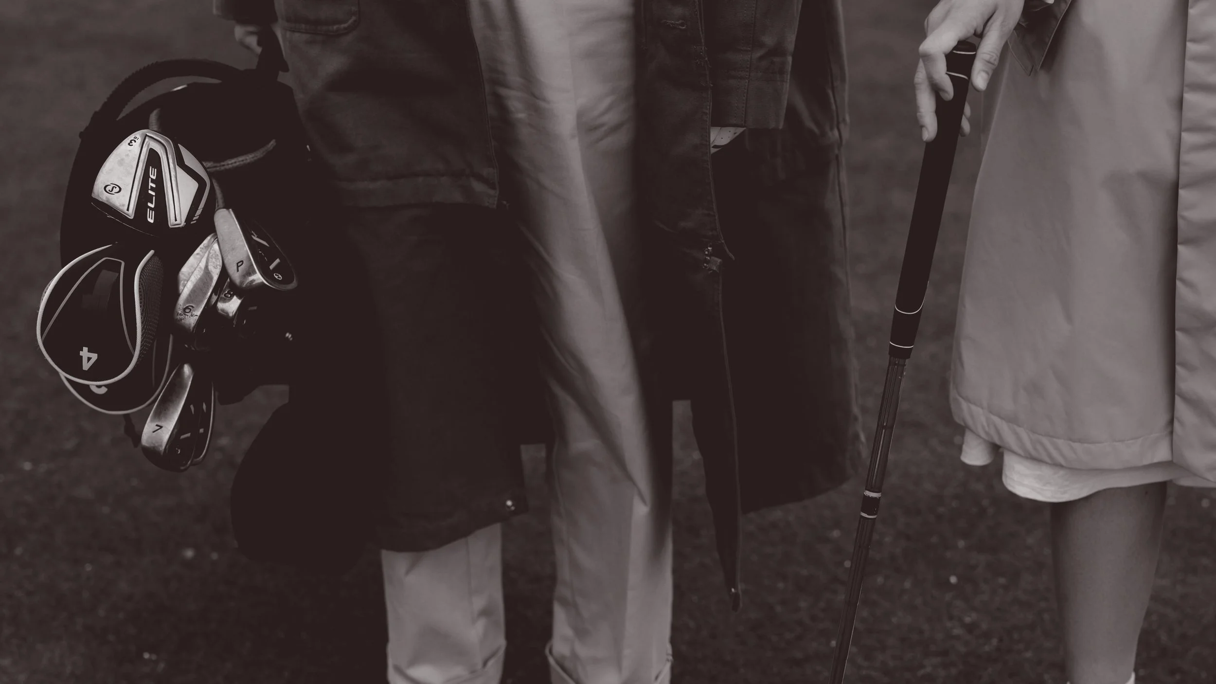 Two people standing on a golf course, one holding a golf club and the other holding a set of golf clubs in a bag.