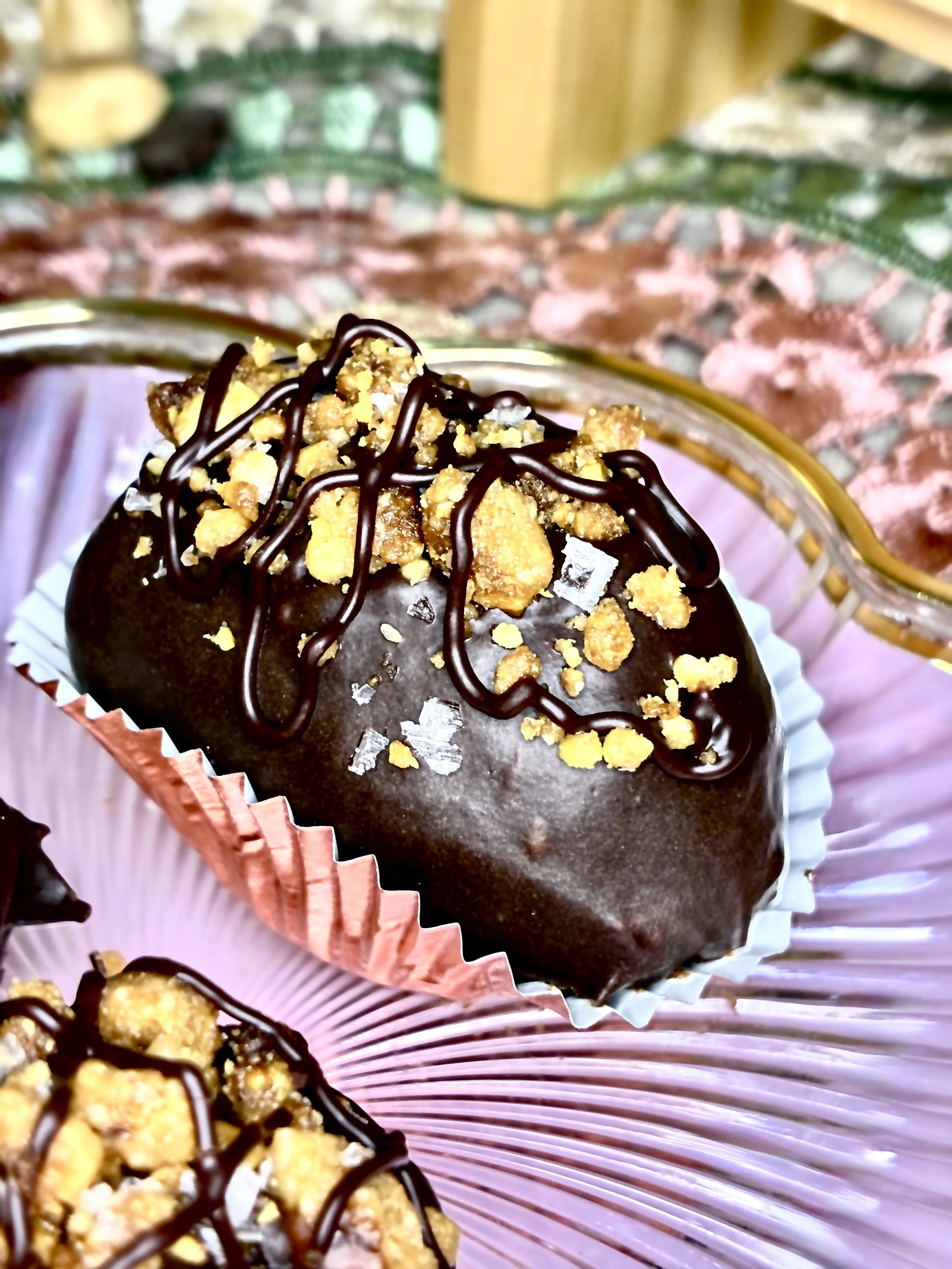 Close-up of a chocolate-covered medjool dates topped with crushed nuts and drizzled with chocolate, placed in a pink paper cup, on a pink and green decorative surface.