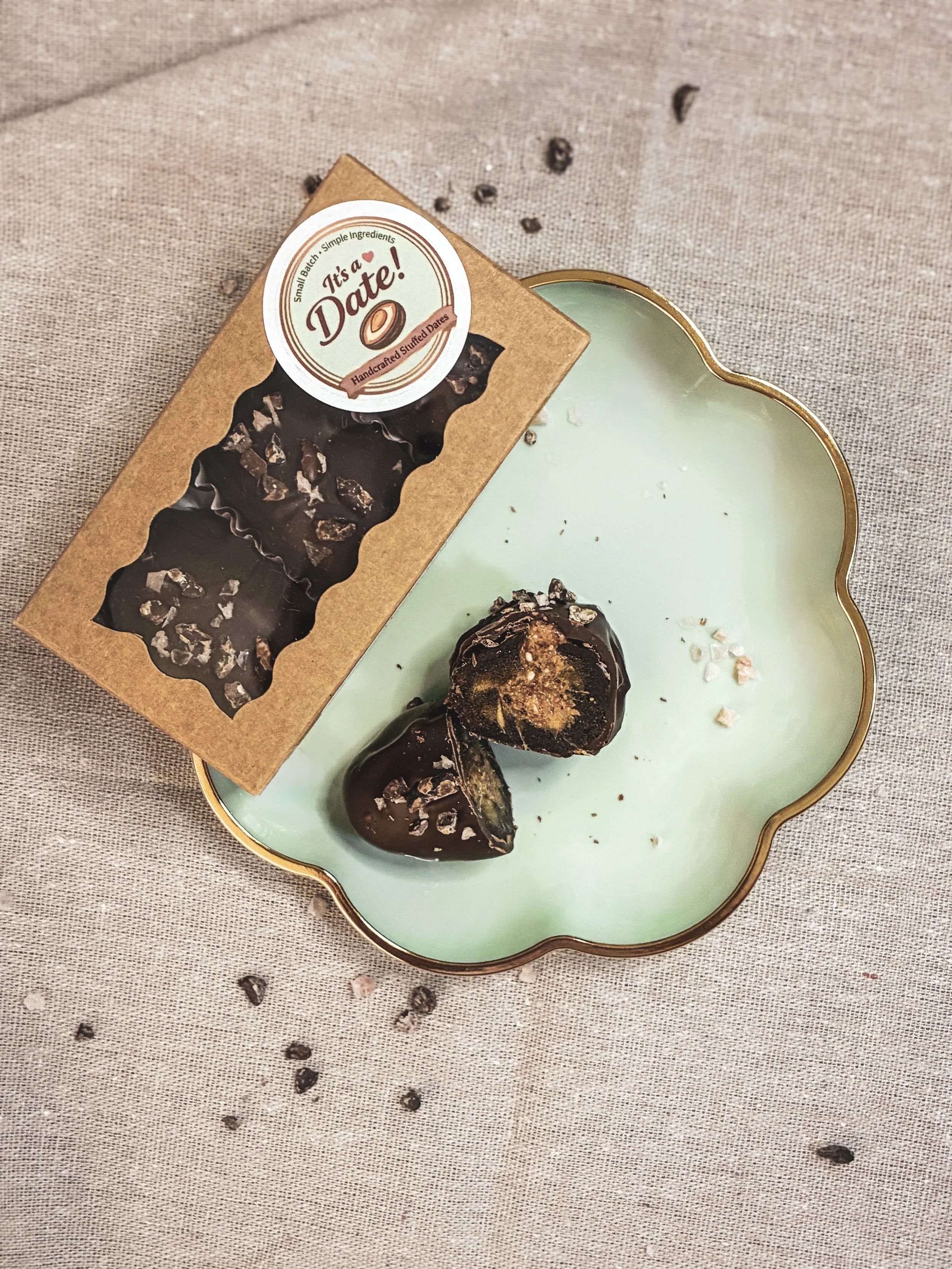 A green decorative plate with gold trim holding two chocolate dipped stuffed dates and some scattered sea salt flakes. A box of handmade stuffed dates is placed next to the plate.