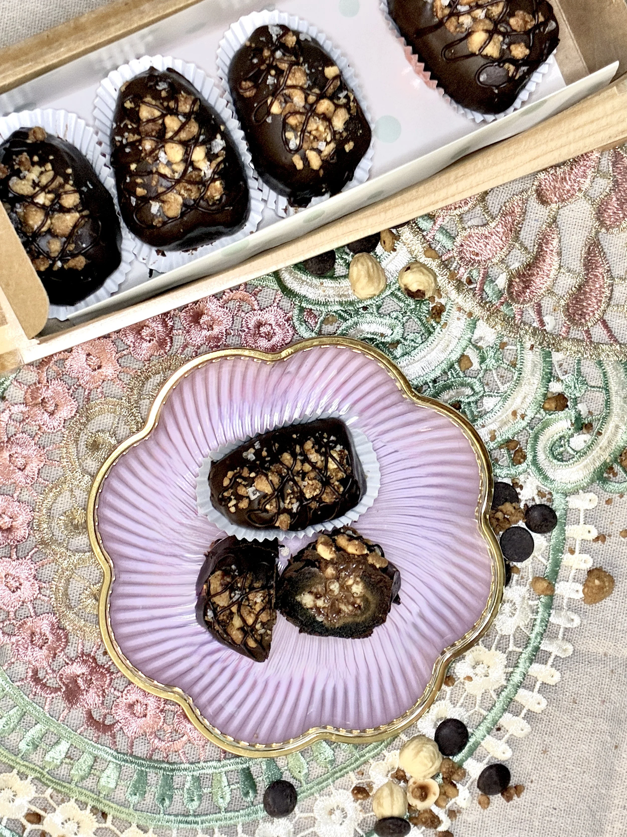 Chocolate-covered medjool dates topped with chopped nuts and drizzled with chocolate sauce on decorative plates placed on embroidered cloth.