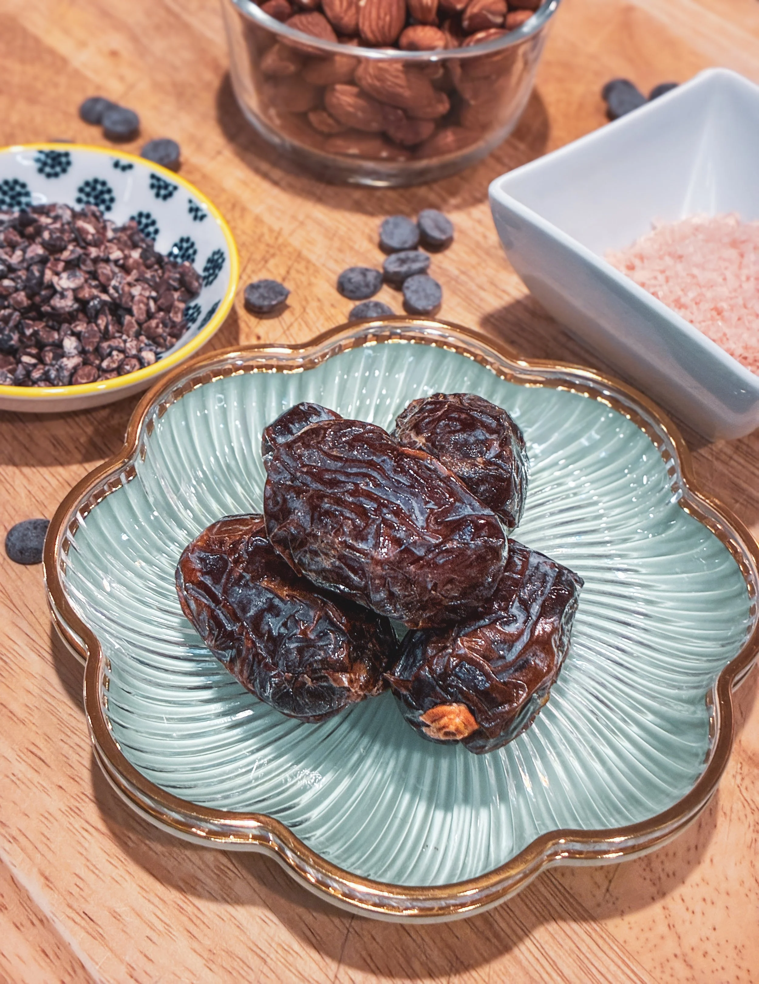 Dried dates in a glass bowl, almonds in a glass container, dried nuts in a yellow and white dish, and chocolate chips in a white dish on a wooden surface.