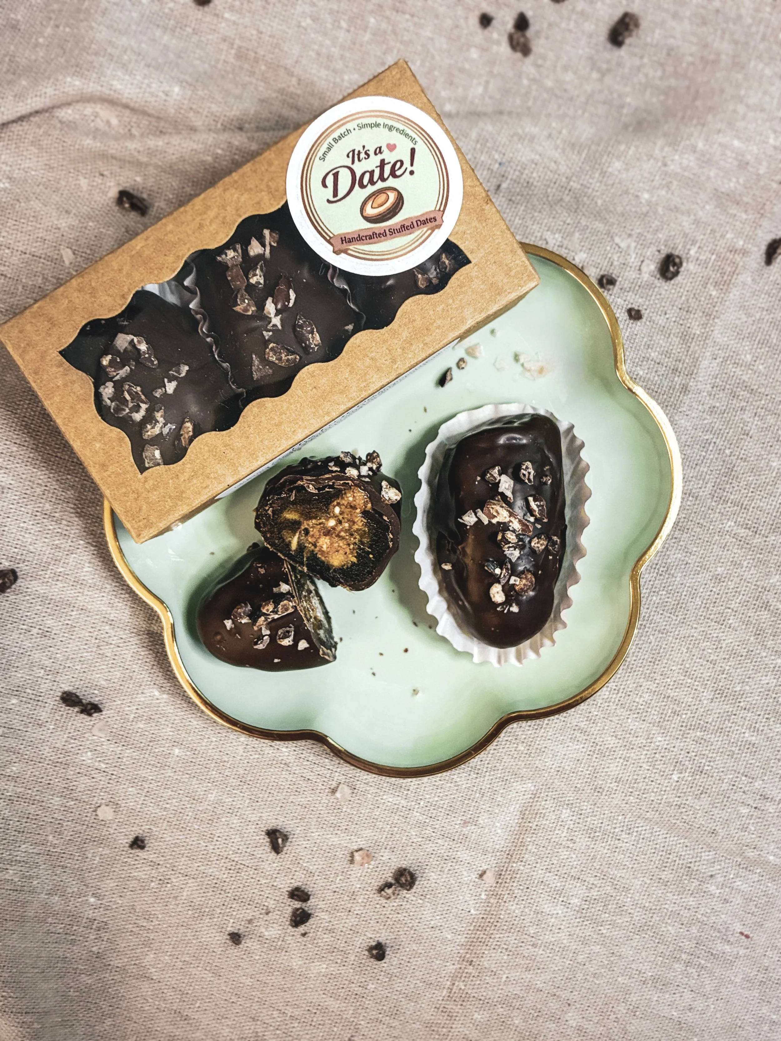 Box of handcrafted stuffed dates with chocolate coating and sprinkled with crushed nuts, placed on a decorative plate with additional stuffed dates.
