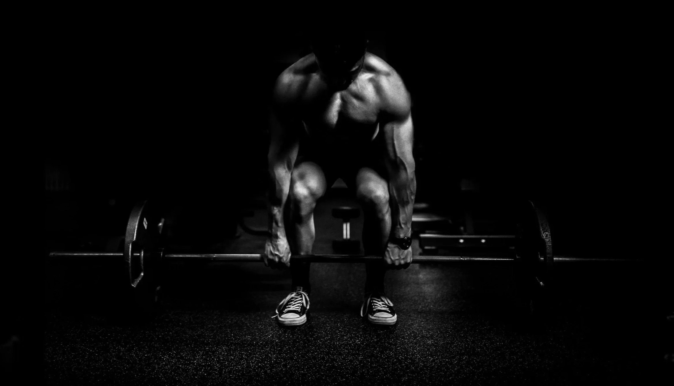 A muscular man in a baseball cap and sneakers is preparing to lift a barbell in a dark gym.