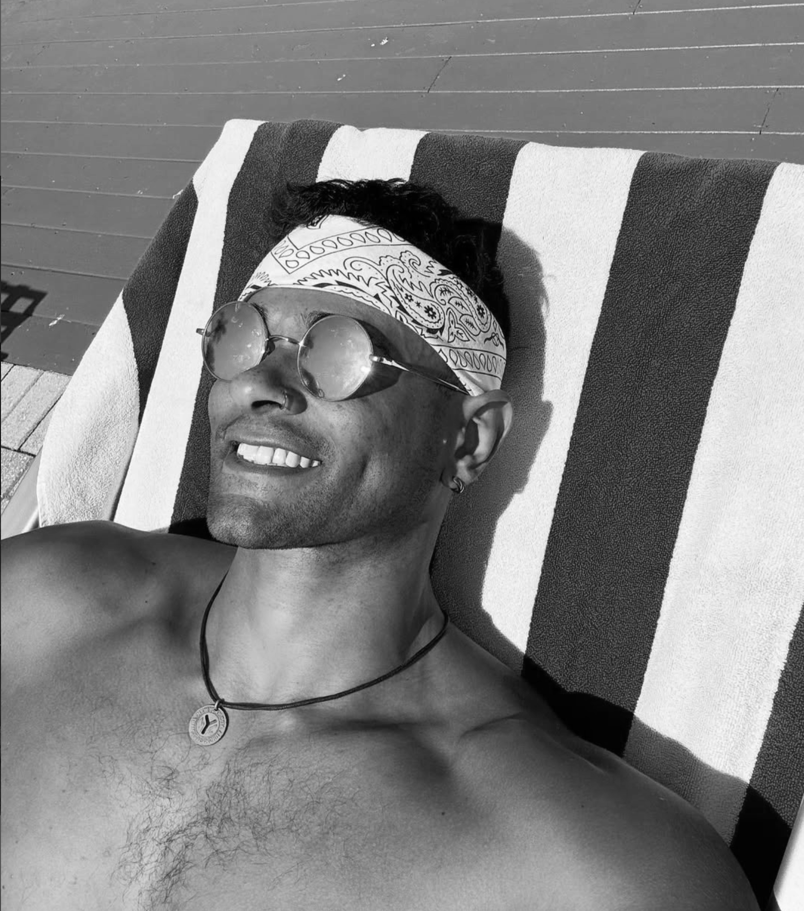 Person lying on a striped towel at the beach, smiling, wearing sunglasses, a bandana, and a necklace.
