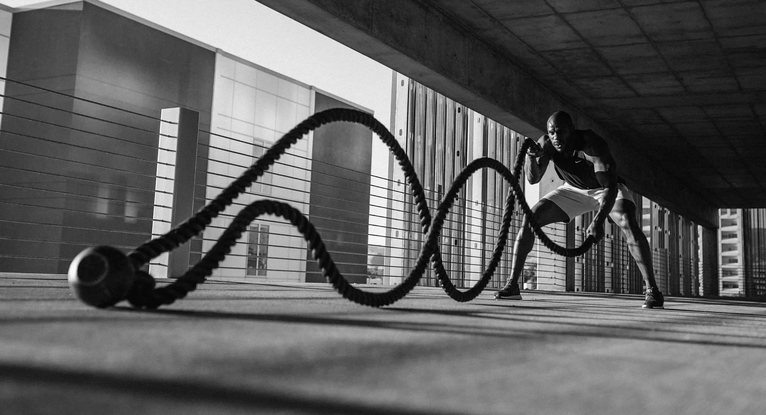 A man exercising outdoors on a balcony, lifting battle ropes. The silhouette shows his muscular build, and he is wearing a sleeveless shirt and shorts. The background reveals tall buildings and a cityscape.