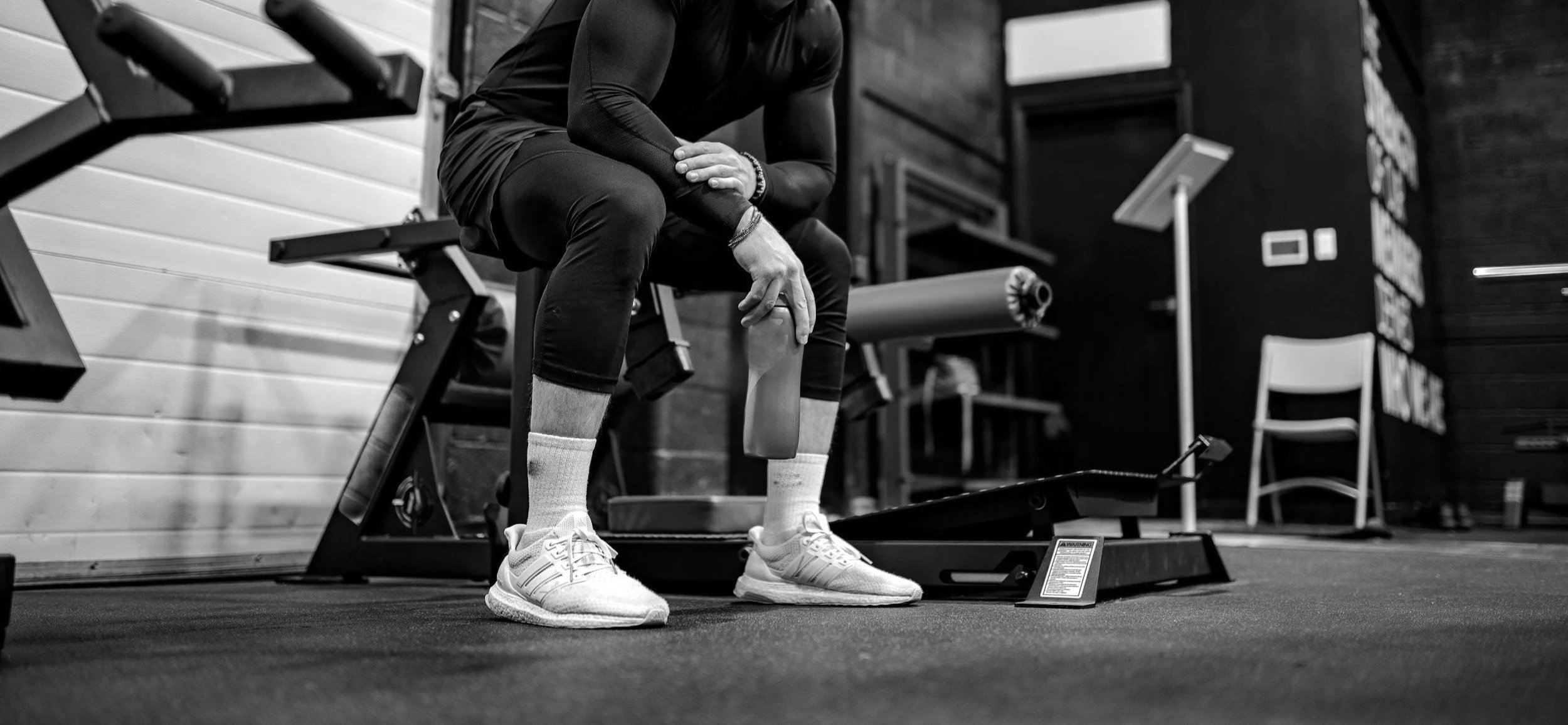A person sitting on a bench in a gym, holding their knee with one hand, wearing athletic clothing and sneakers.
