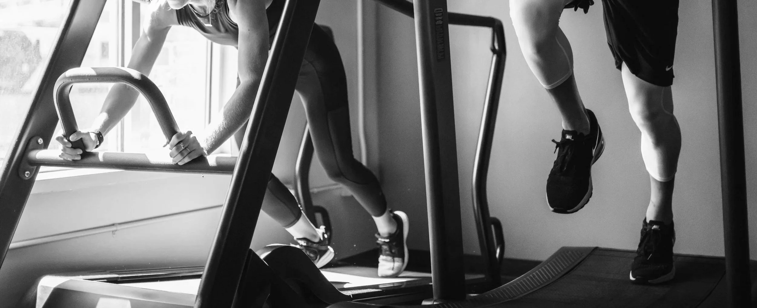 Two people are exercising on cardio machines in a gym, with one person walking on a treadmill and another using an elliptical trainer, both wearing workout clothes and sneakers.