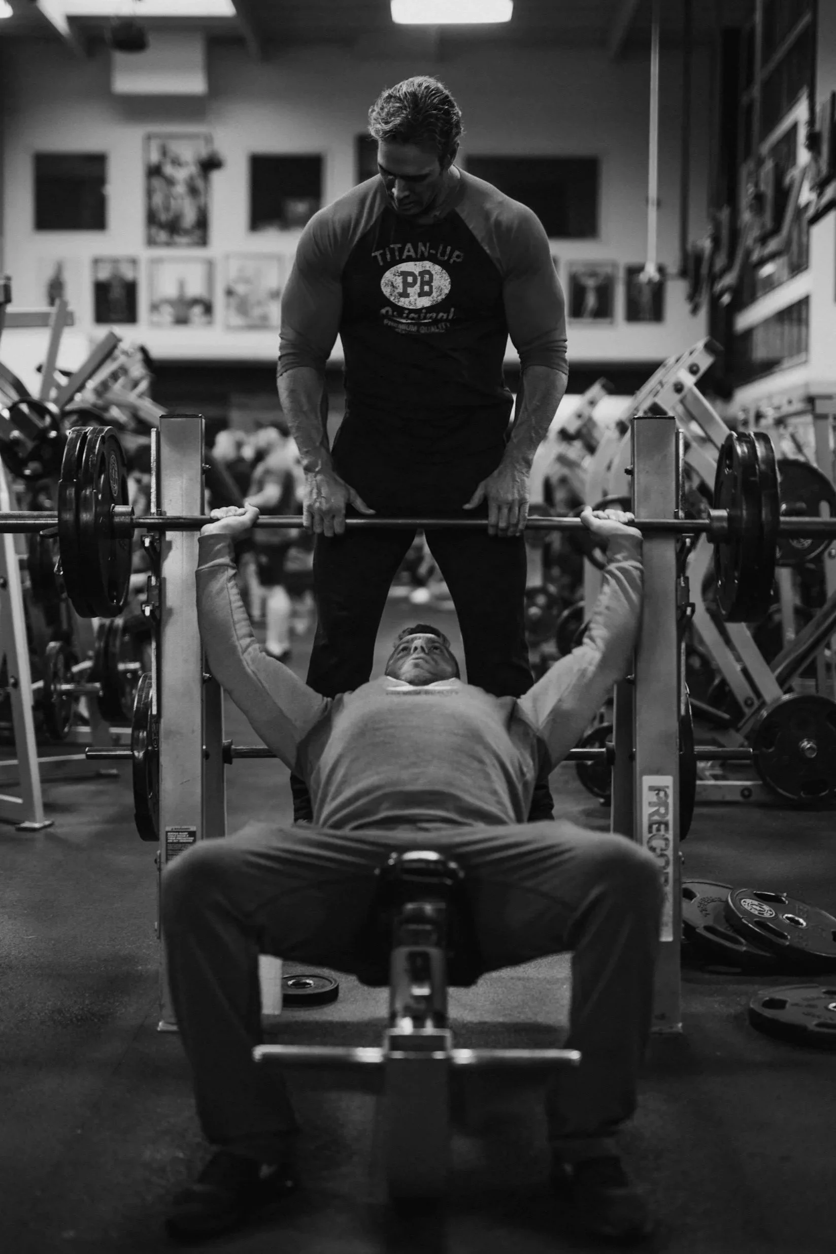A man spotting another man lifting a barbell during a workout in a gym.