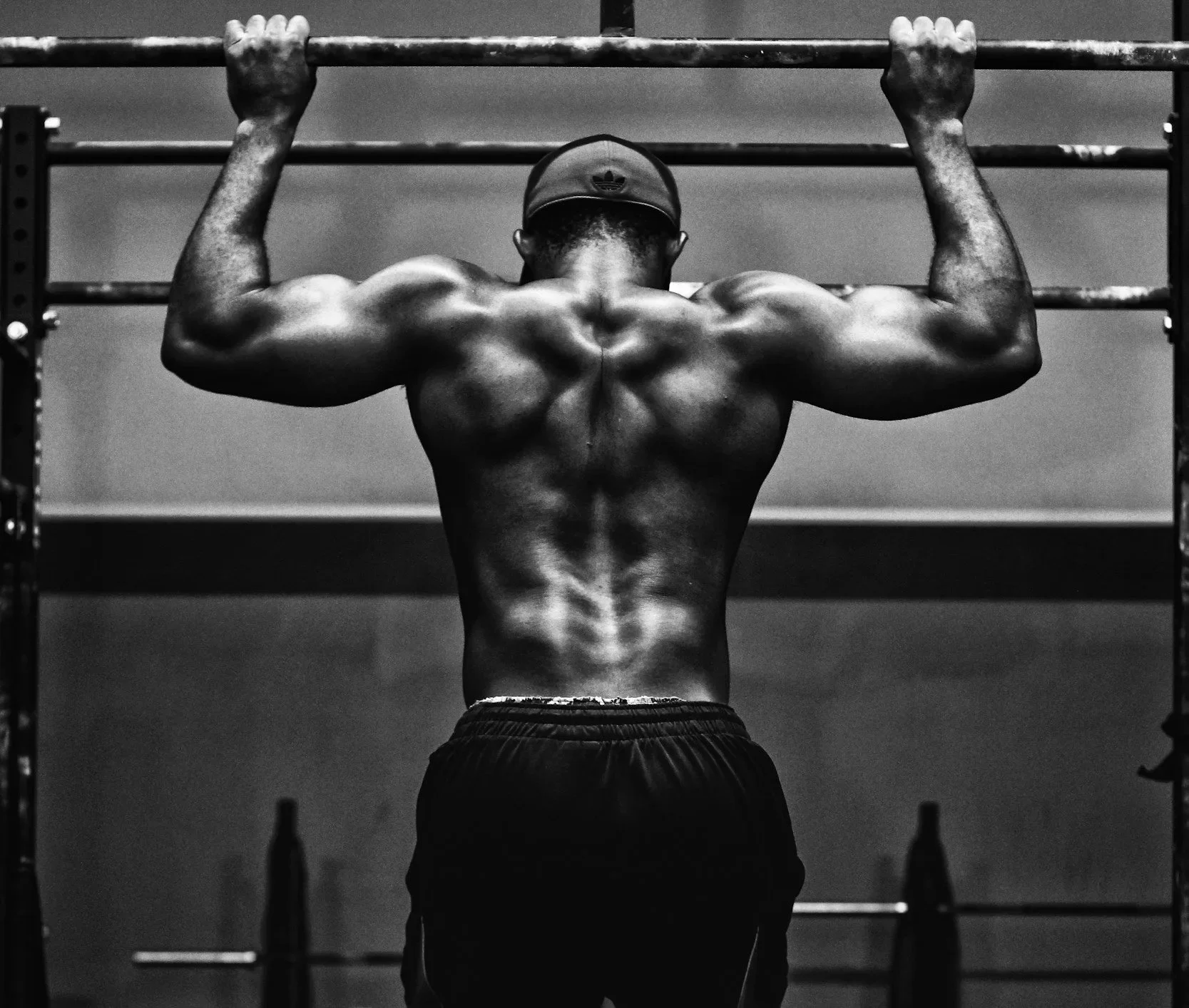 A shirtless man with a cap, seen from behind, doing pull-ups on a bar in a gym.