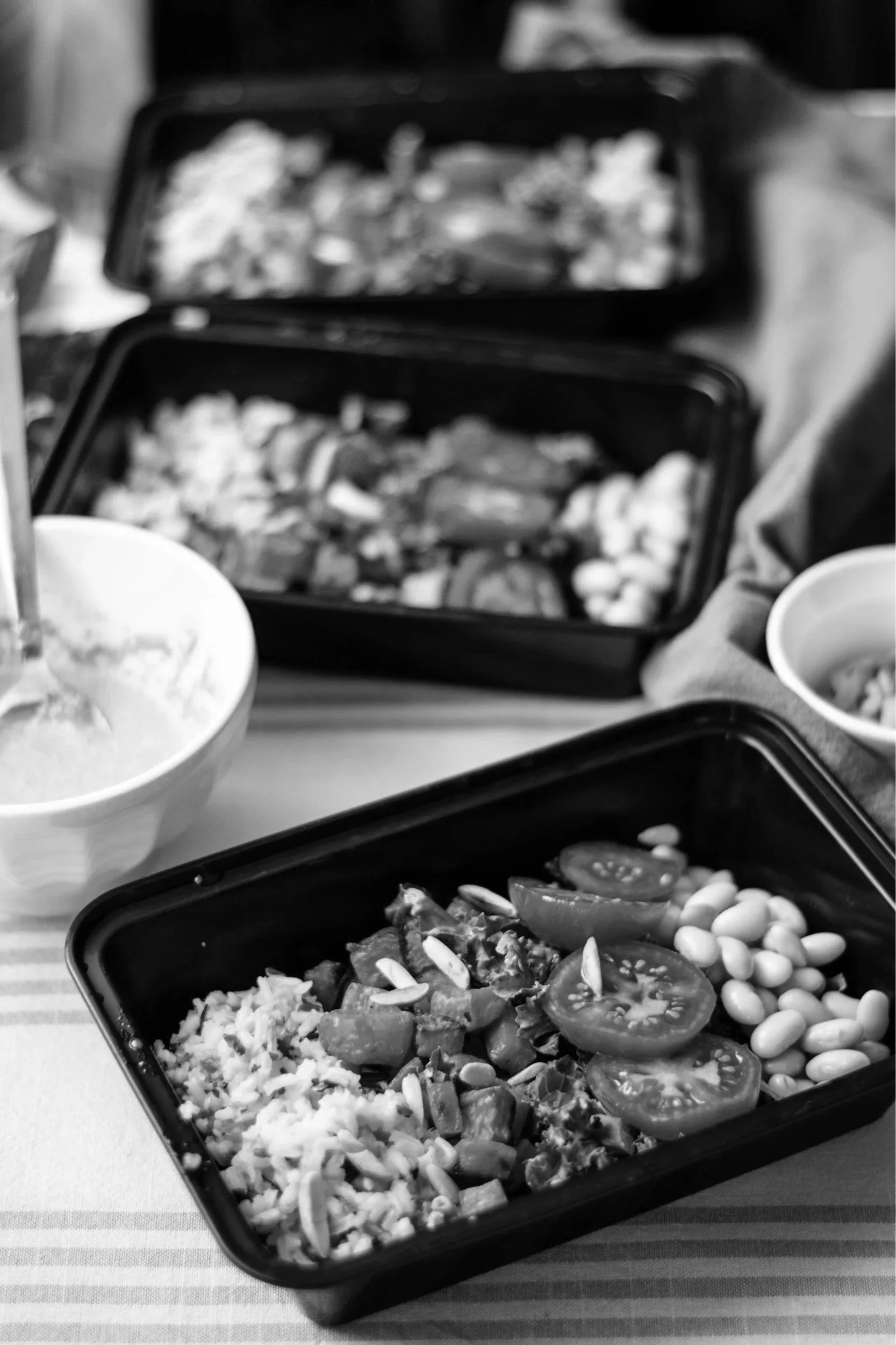 Meal containers with sliced tomatoes, leafy greens, and beans, along with bowls of salad and utensils on a striped tablecloth.
