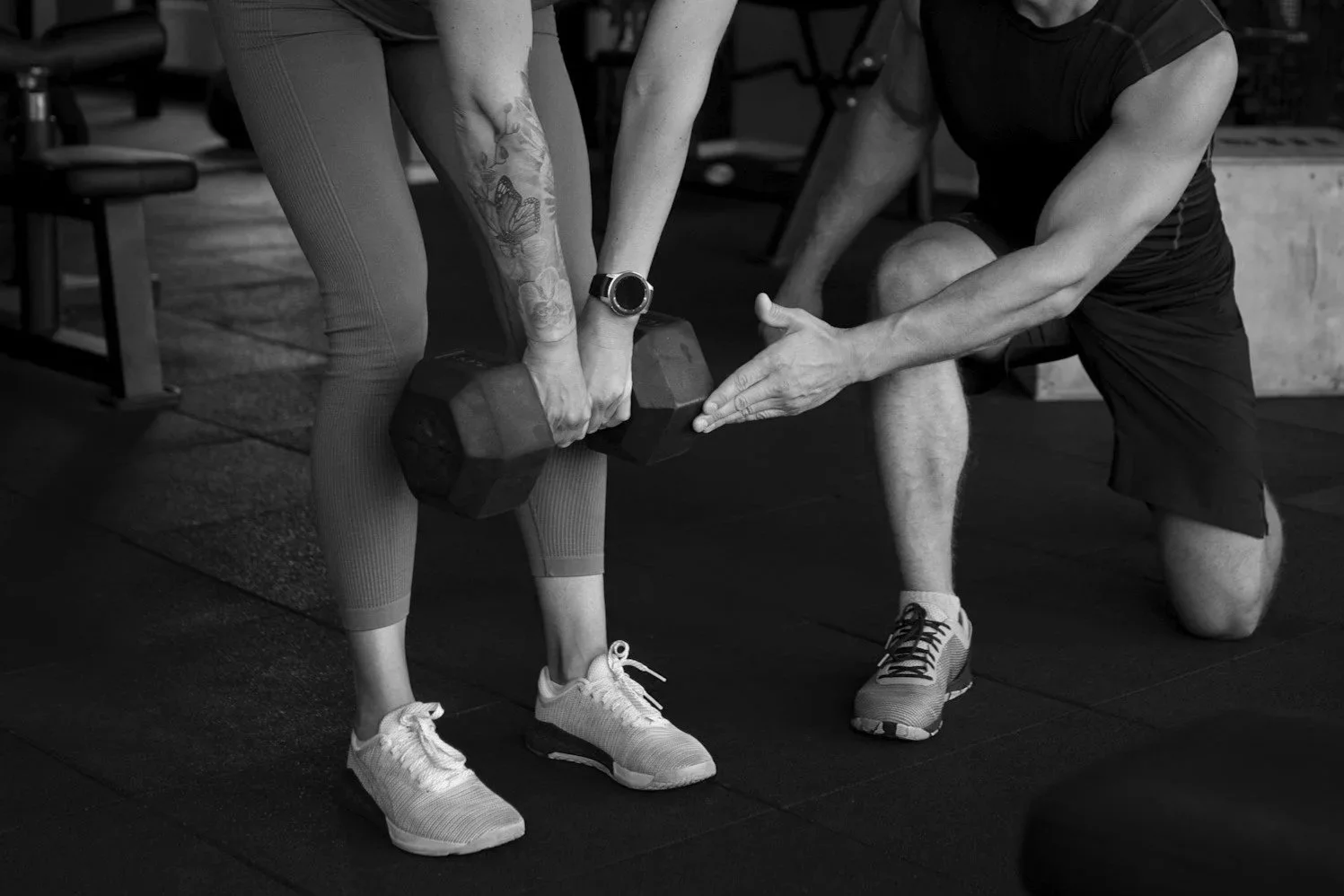 A woman lifting a dumbbell with assistance from a trainer in a gym. The trainer is kneeling, supporting the woman's arm, while she stands with her legs apart and exercises with the weight.