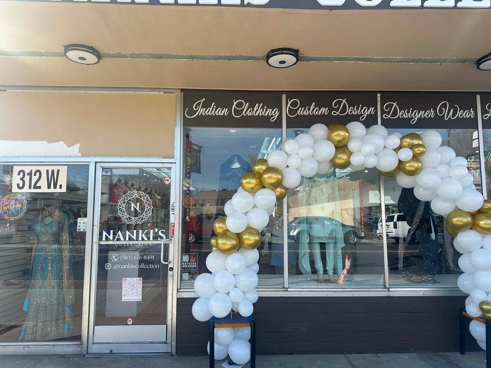 Storefront of Nanki's Collection with a balloon arch made of white and gold balloons, mannequins inside displaying Indian clothing, and signage advertising Indian clothing, custom design, and designer wear.