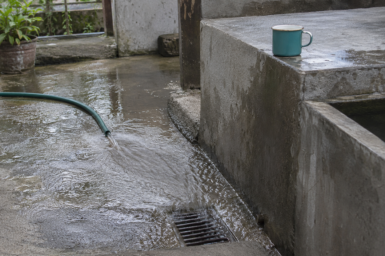 Water flows from a green hose toward a drain in a concrete courtyard, with a teal cup on a raised ledge.