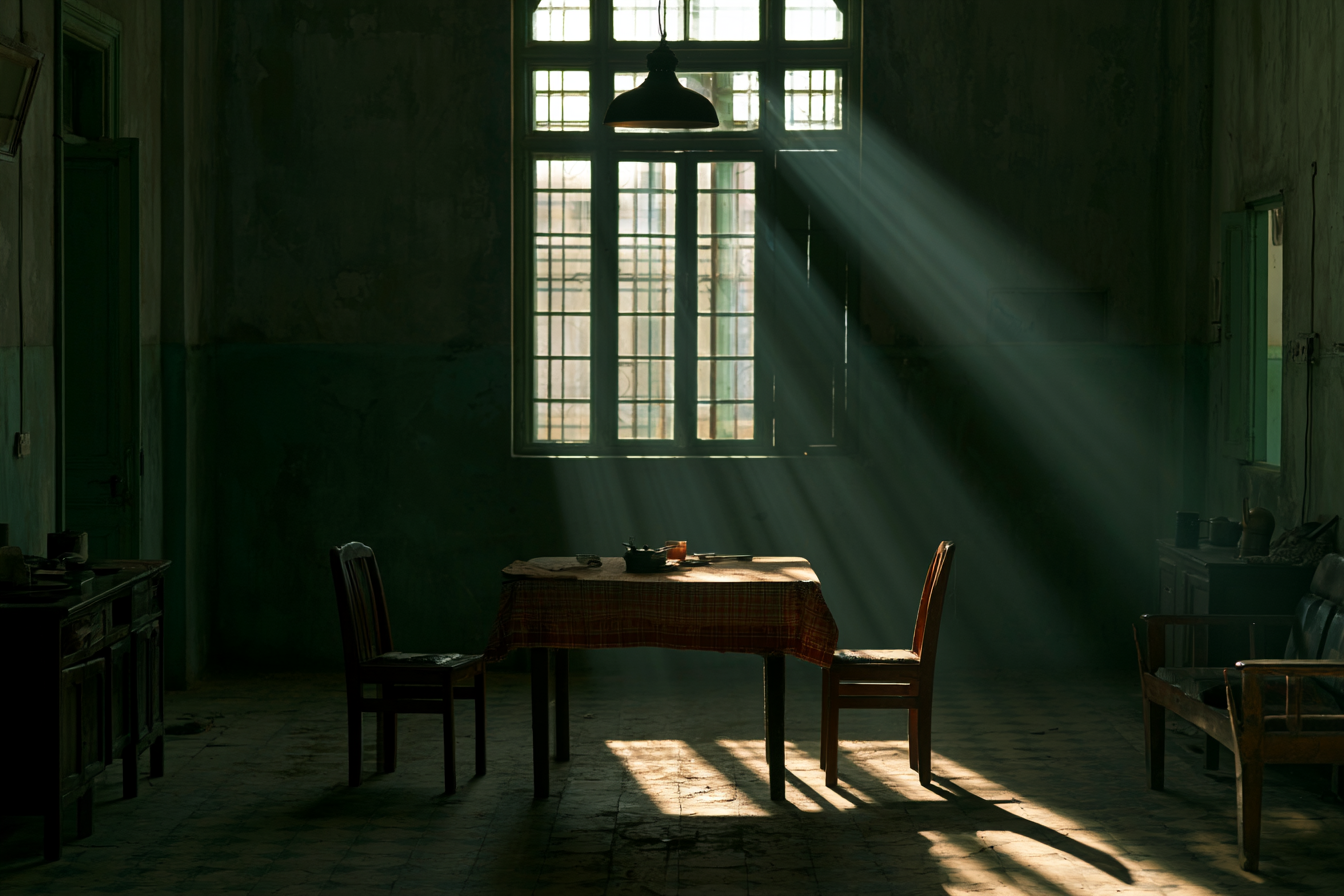 An empty room with a tall bright window, a table, two facing chairs and bands of light across the floor.