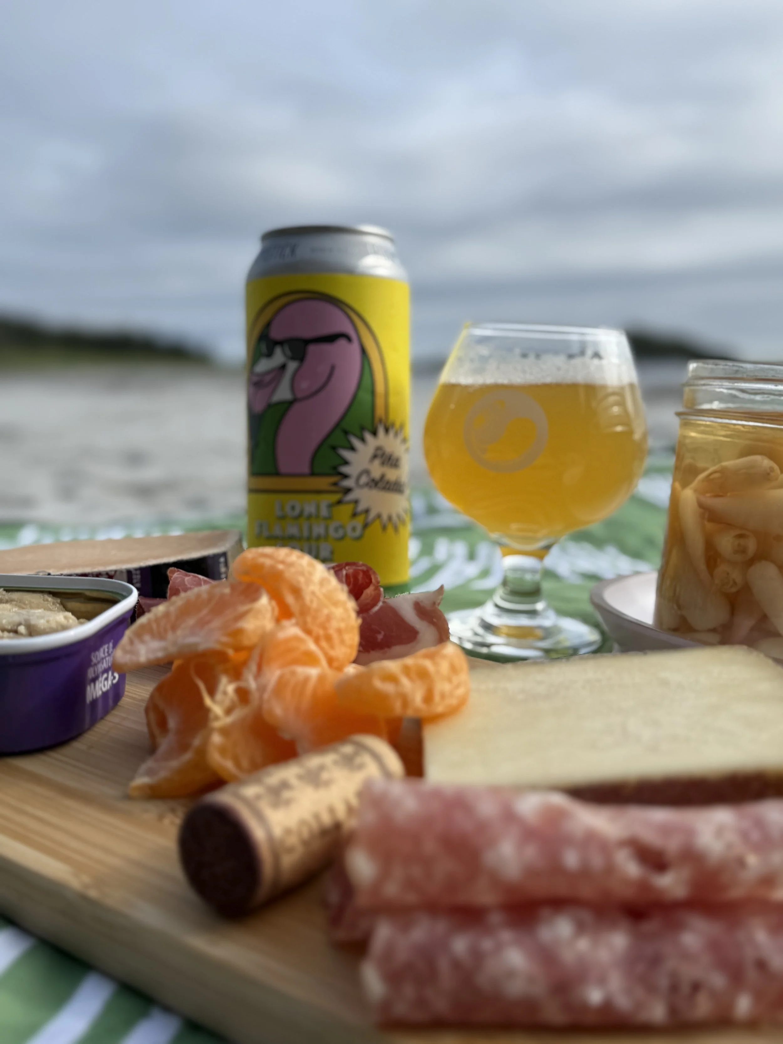 A picnic scene at the beach with food and drinks, including slices of orange, cured meats, a can of La Panzona Flamingo beer, and glasses of beer, with a cloudy sky in the background.