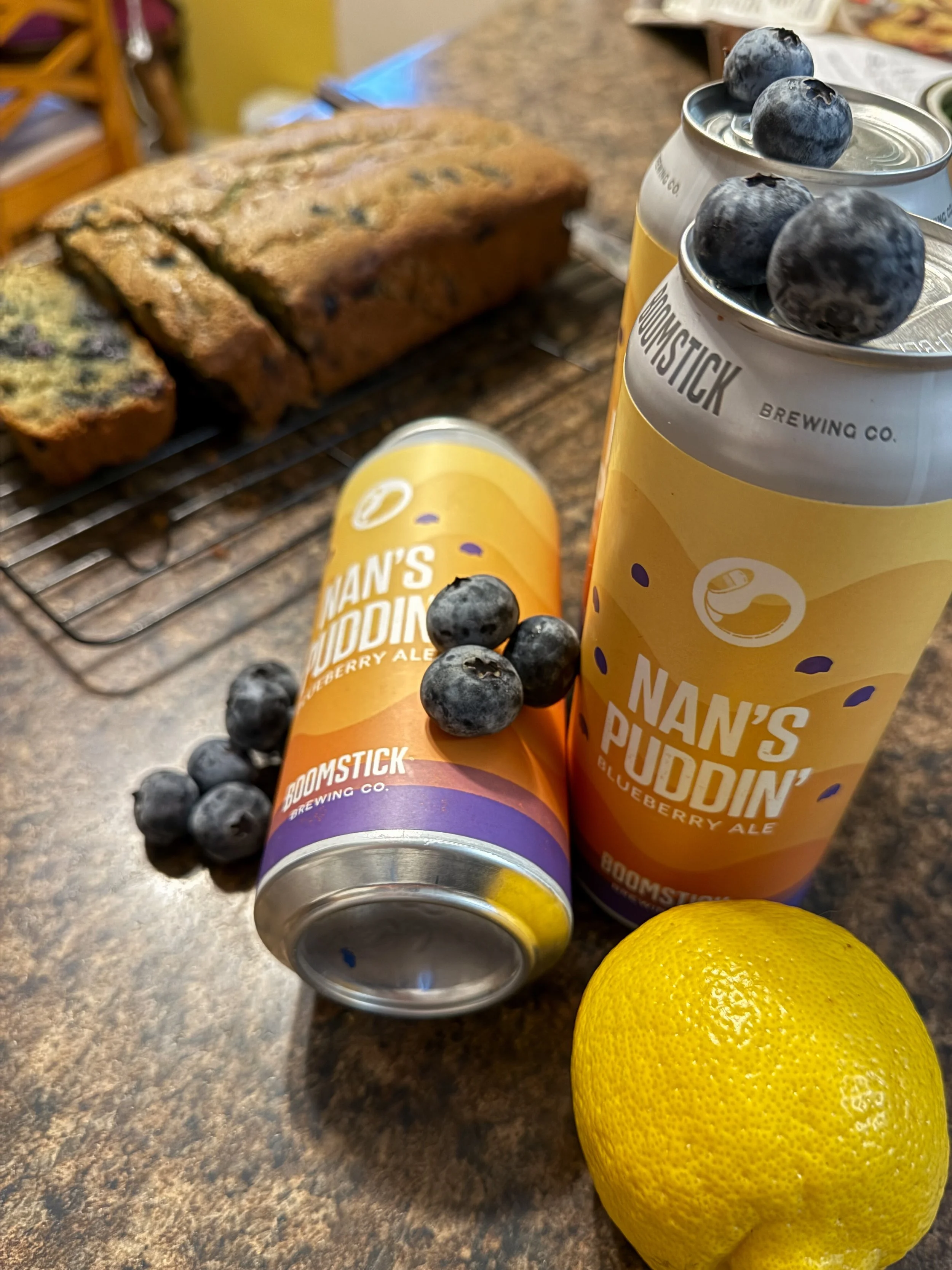 Two cans of Boomstick Brewing blueberry ale, two fresh lemons, and a blueberry cake bread on a kitchen countertop.