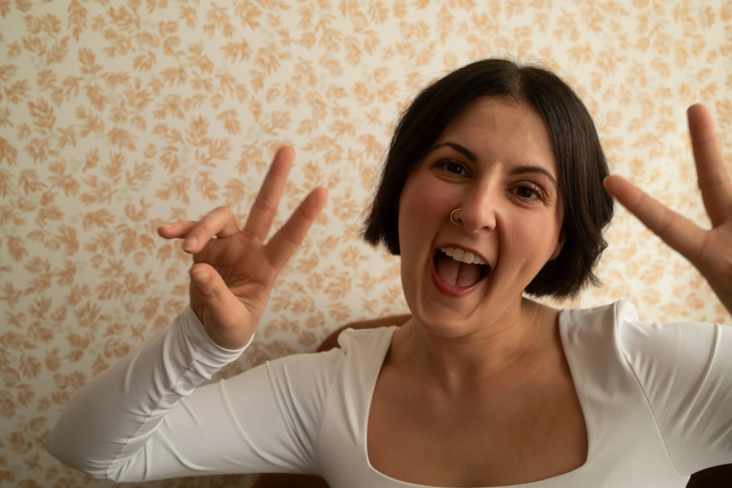 A woman with short dark hair, wearing a white top, is smiling and making peace signs with both hands in front of a beige floral patterned wall.
