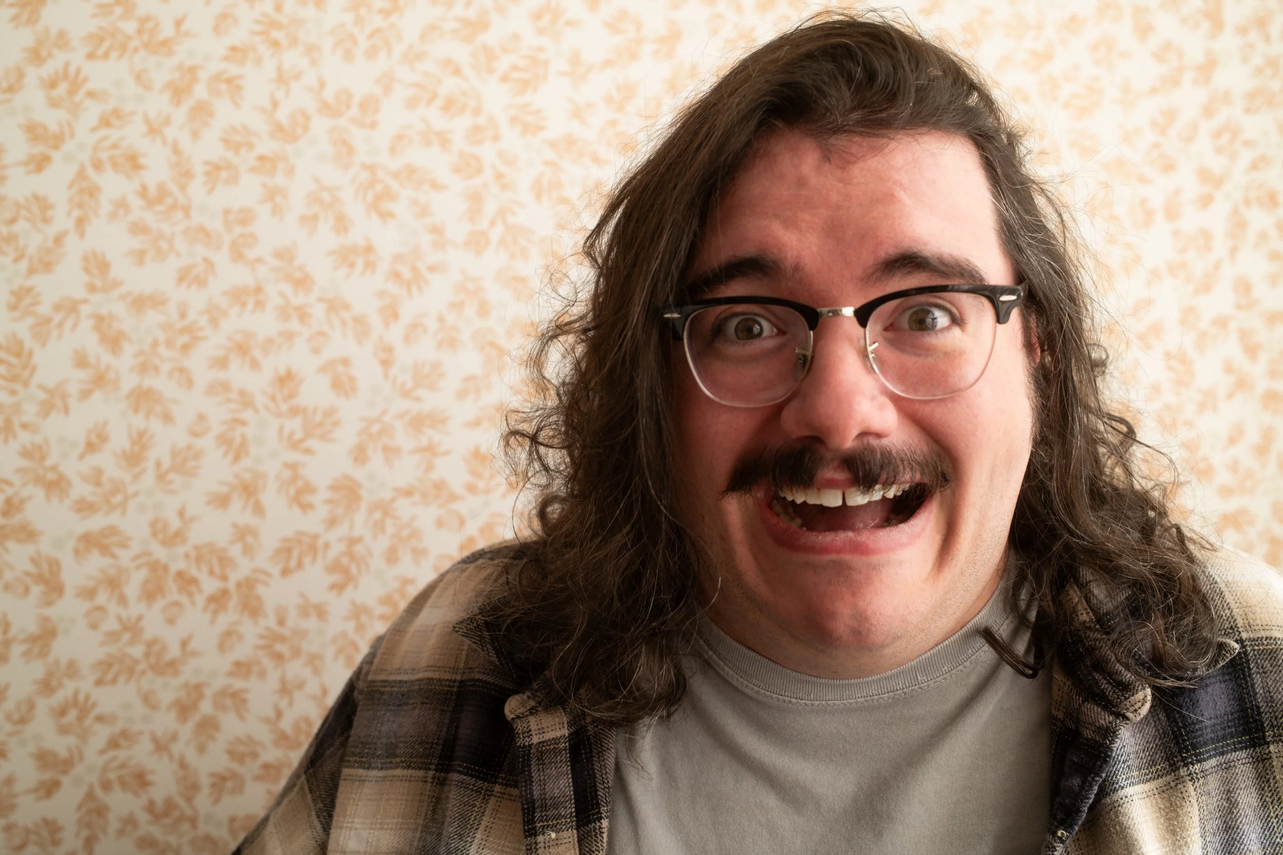 A man with long curly brown hair, glasses, and a mustache, smiling widely showing his teeth. He's wearing a gray t-shirt and a plaid shirt, standing in front of a light-colored wallpaper with orange floral patterns.