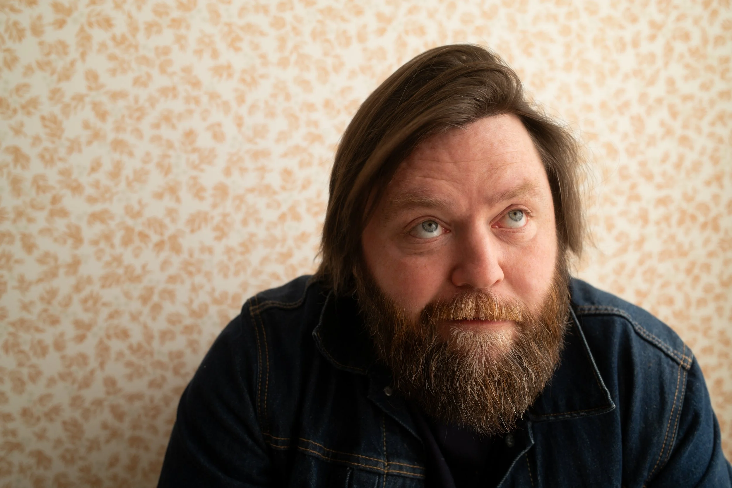 A man with light skin, bluish eyes, brown hair, and a full beard, wearing a denim jacket, looking upwards, in front of a beige patterned wall.