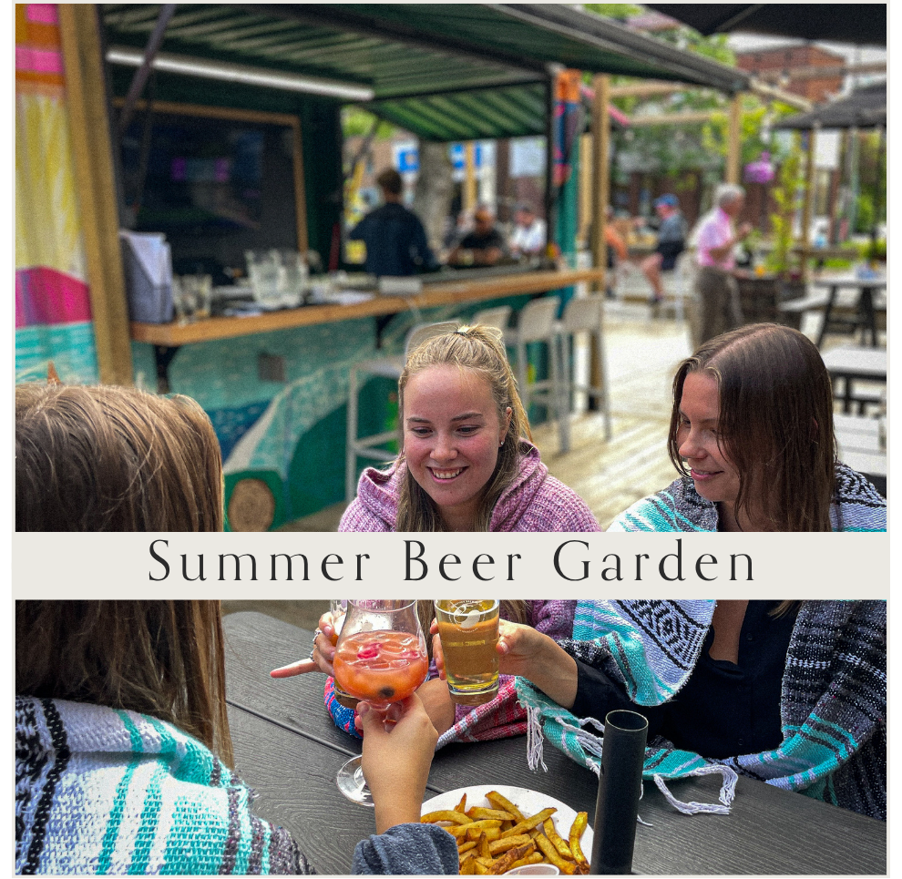 Three young women toasting with drinks at an outdoor summer beer garden, with food on the table, surrounded by other people and outdoor seating.