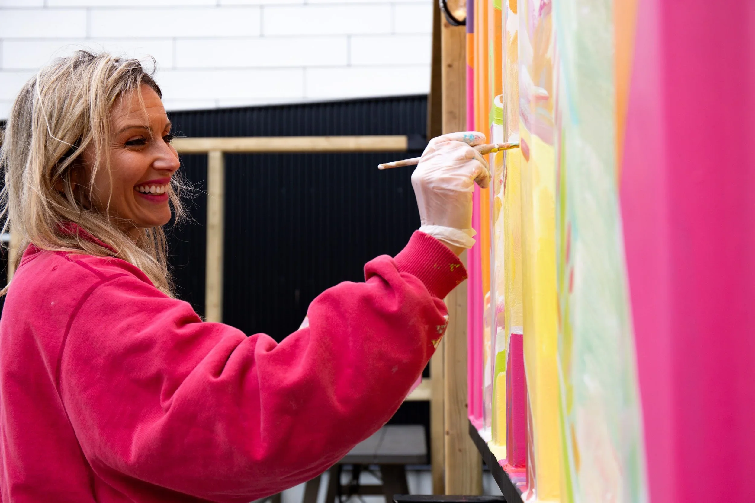 A woman with blonde hair, wearing a red sweatshirt, smiling while painting on a colorful wall with pink, yellow, and other pastel shades.