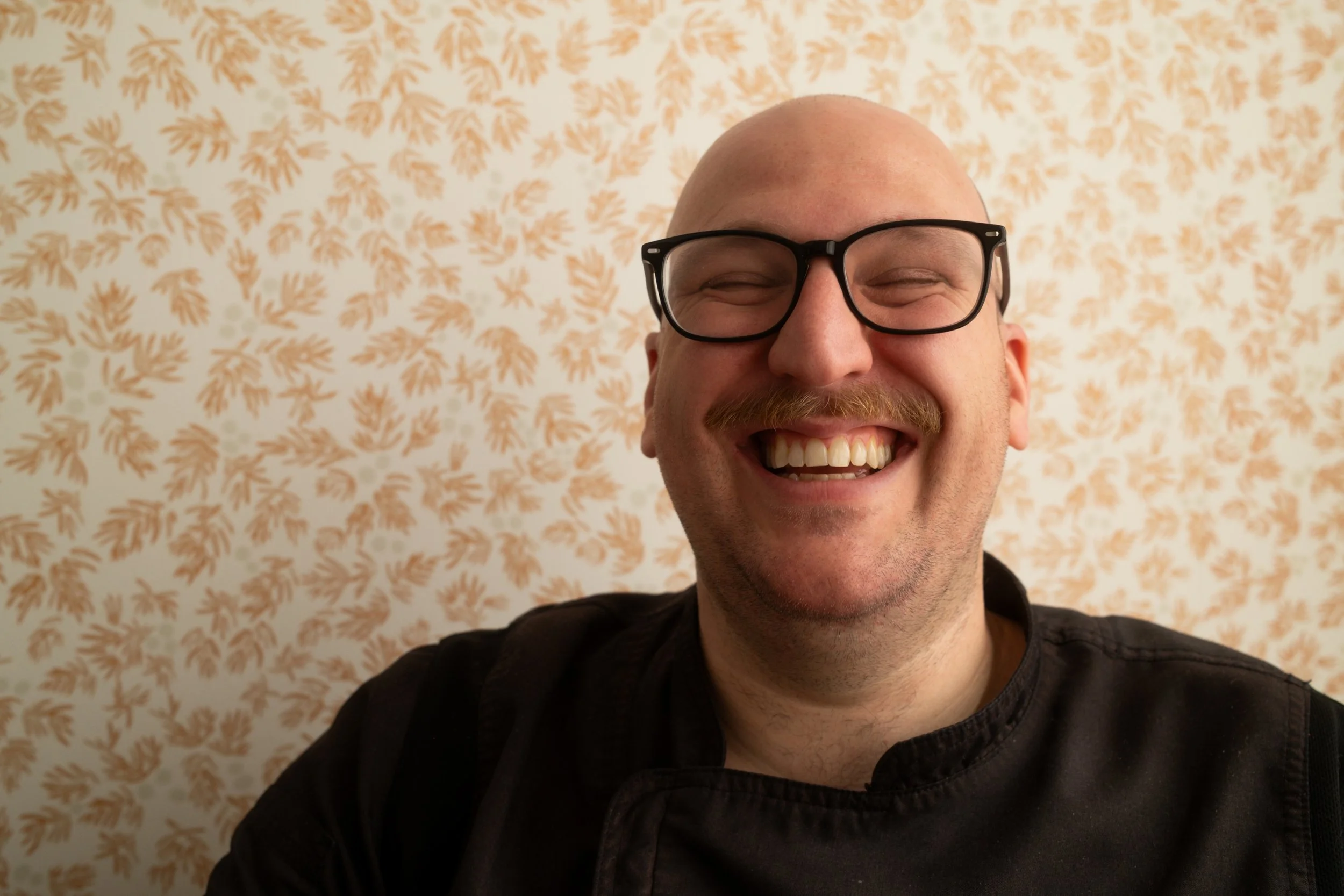 A smiling man with glasses and a mustache taking a selfie in front of a patterned wallpaper with orange botanical designs.