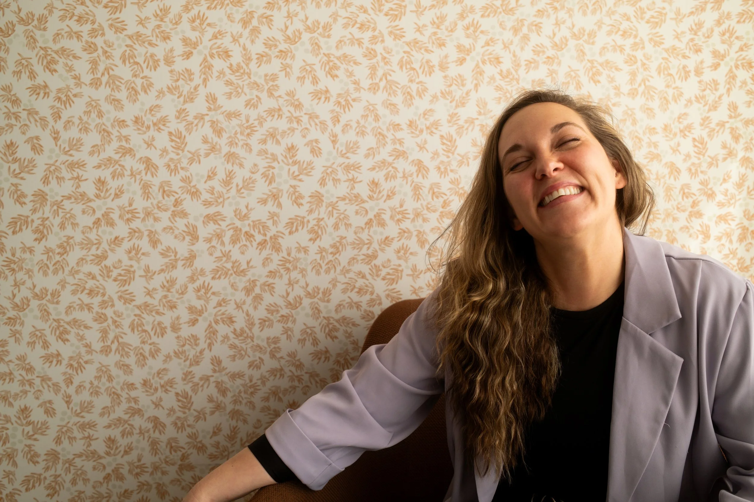 A woman with long curly hair smiling and sitting on a brown chair in front of a beige patterned wallpaper.