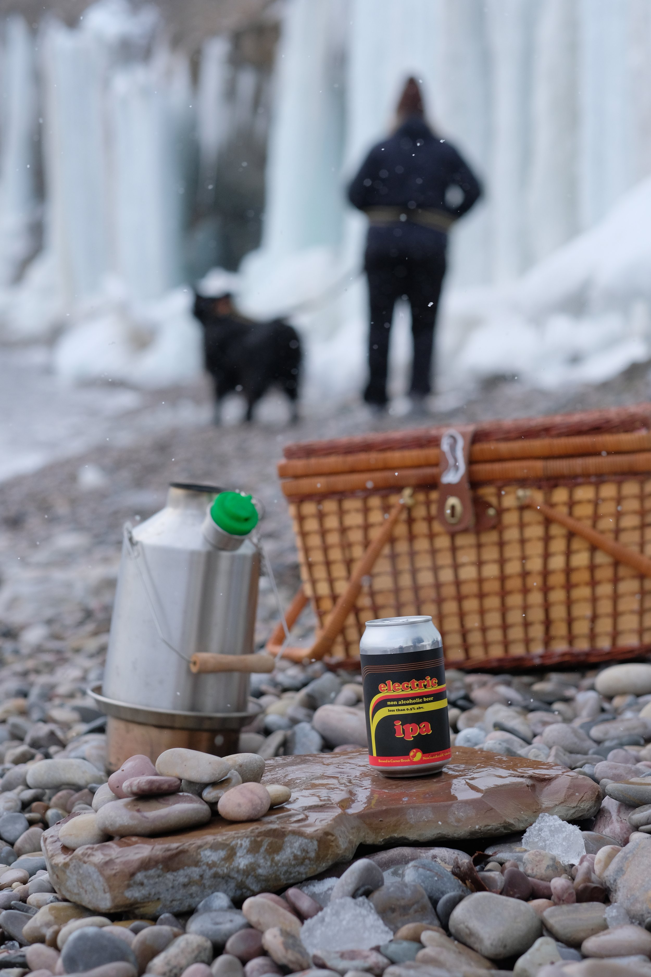 A close-up of a rocky beach with miscellaneous items including a can of electric IPA non-alcoholic beer, a thermos with a green lid, a wicker basket, and a flat stone. In the background, a person and a dog stand near a waterfall or ice formations, ou