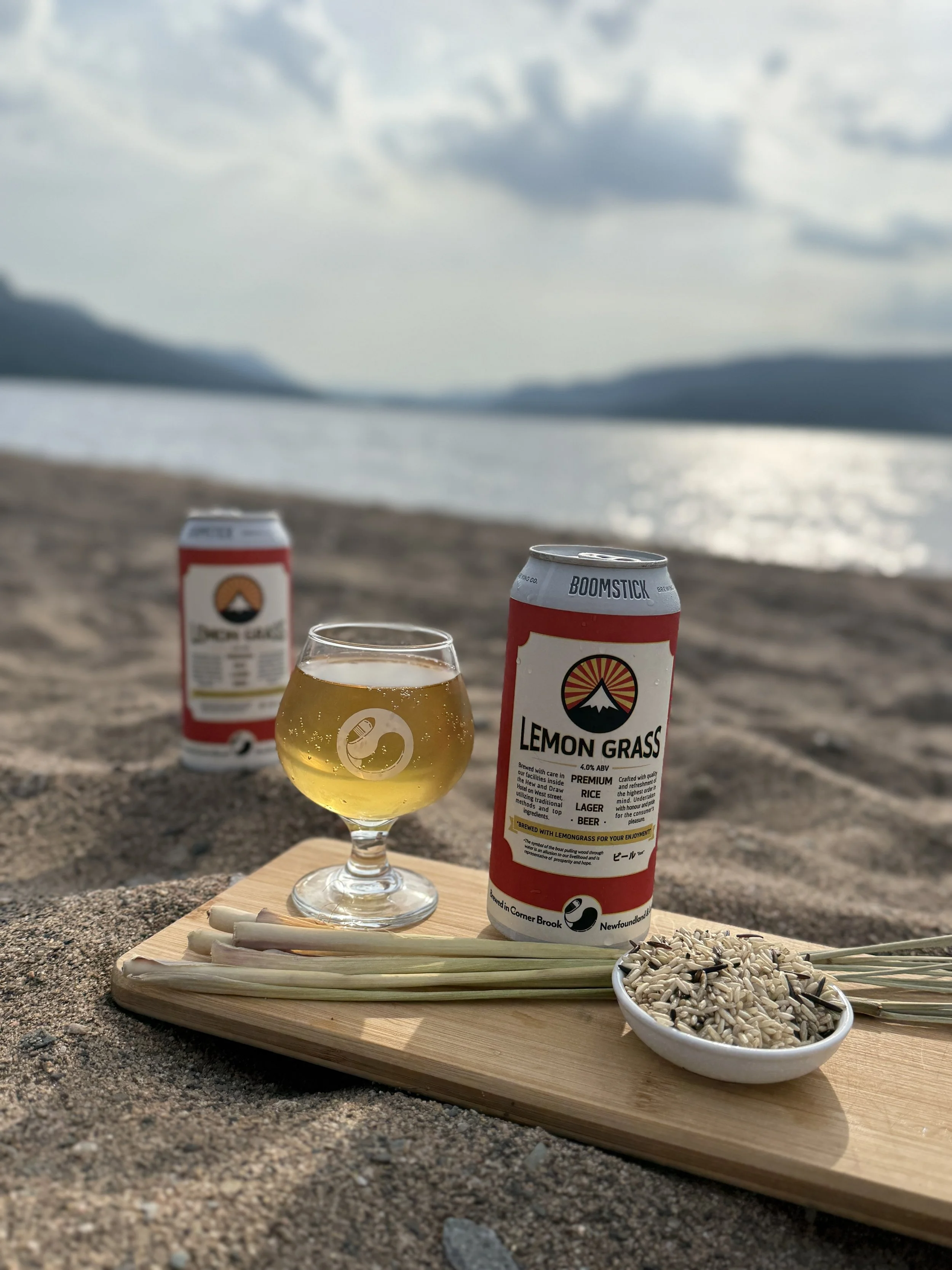 A scene at the beach with a wooden tray holding a glass of beer, a bowl of sunflower seeds, and chopsticks, with two cans of Lemon Grass beer and two cans of Jack's Lager in the background, mountains, water, and cloudy sky in the distance.