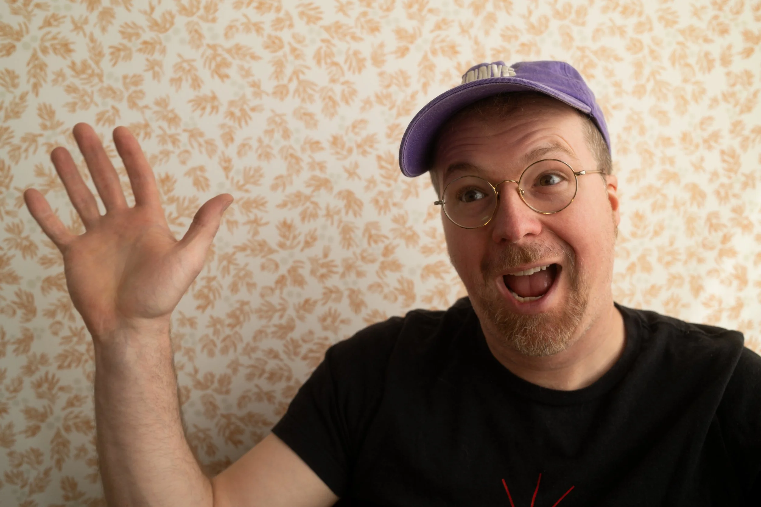 A man with glasses and a purple cap smiling and waving at the camera, standing in front of a patterned wallpaper.
